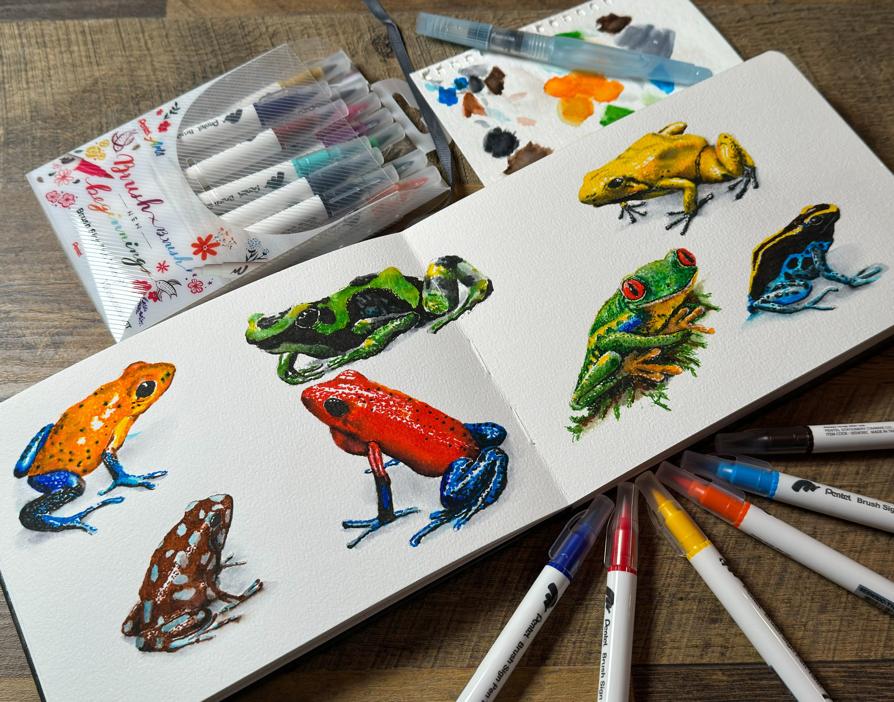 A collection of marker drawings of various colorful frogs on a textured white canvas, surrounded by markers and water brushes, with a piece of paper showing paint smudges and mixing areas in the background, all laid out on a wooden surface.