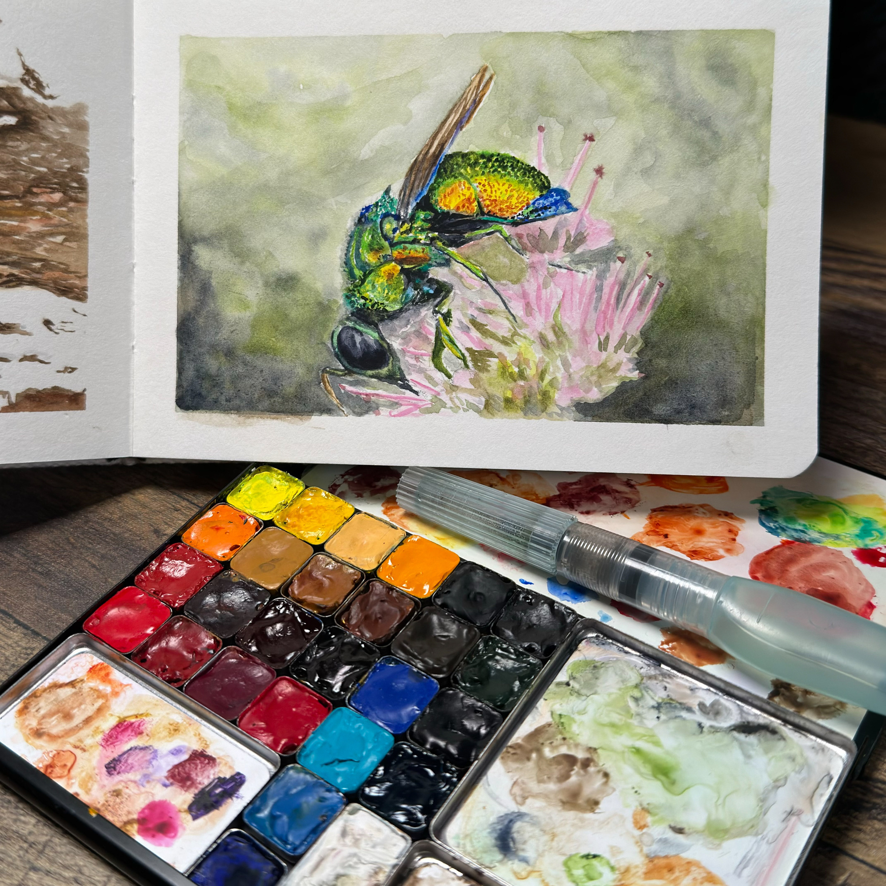 A watercolor painting of an emerald cuckoo wasp with its shimmering green and blue hues, perched delicately on a cluster of pink flowers. Below the artwork is a well-used palette brimming with a rainbow of watercolor paints, accompanied by a water brush pen, all resting on a rustic wooden surface.