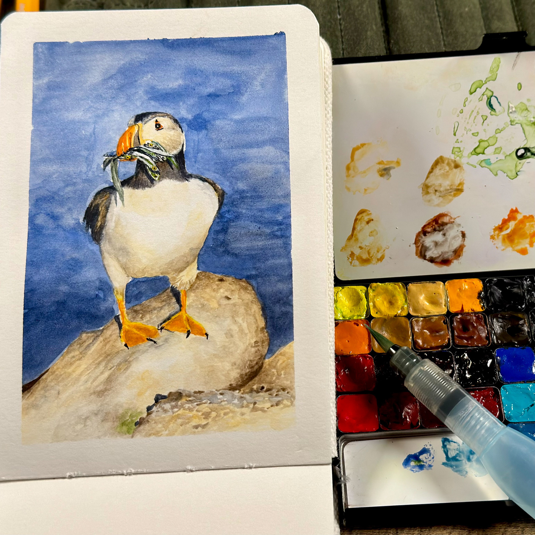 Watercolor painting of a puffin standing on a rock, holding fish in its beak, displayed alongside a watercolor palette and a Pentel water brush.