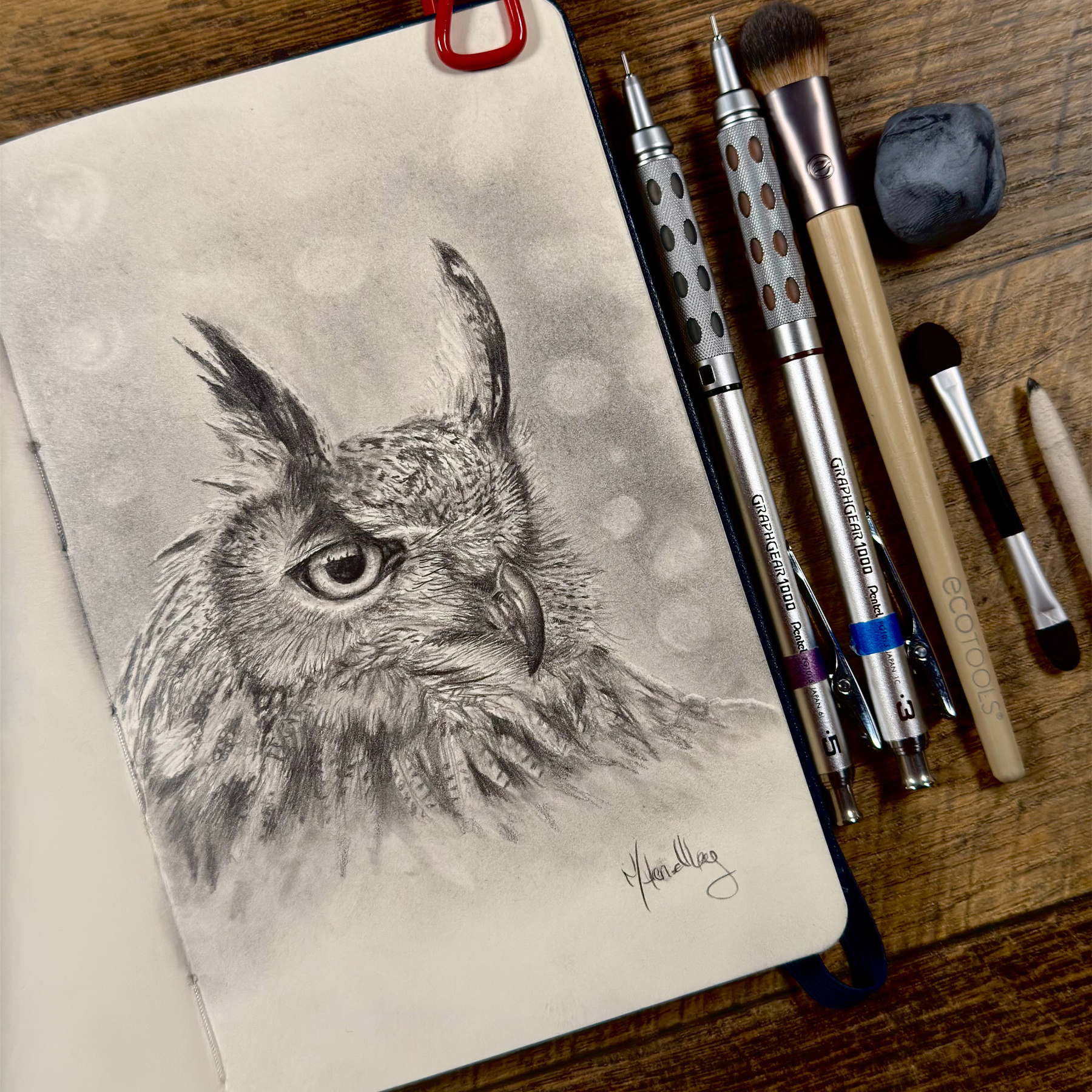 A detailed graphite drawing of an Eurasian eagle owl in a sketchbook. Tools, including graphite pencils, blending stumps, brushes, and a kneaded eraser, are displayed beside it on a wooden surface.