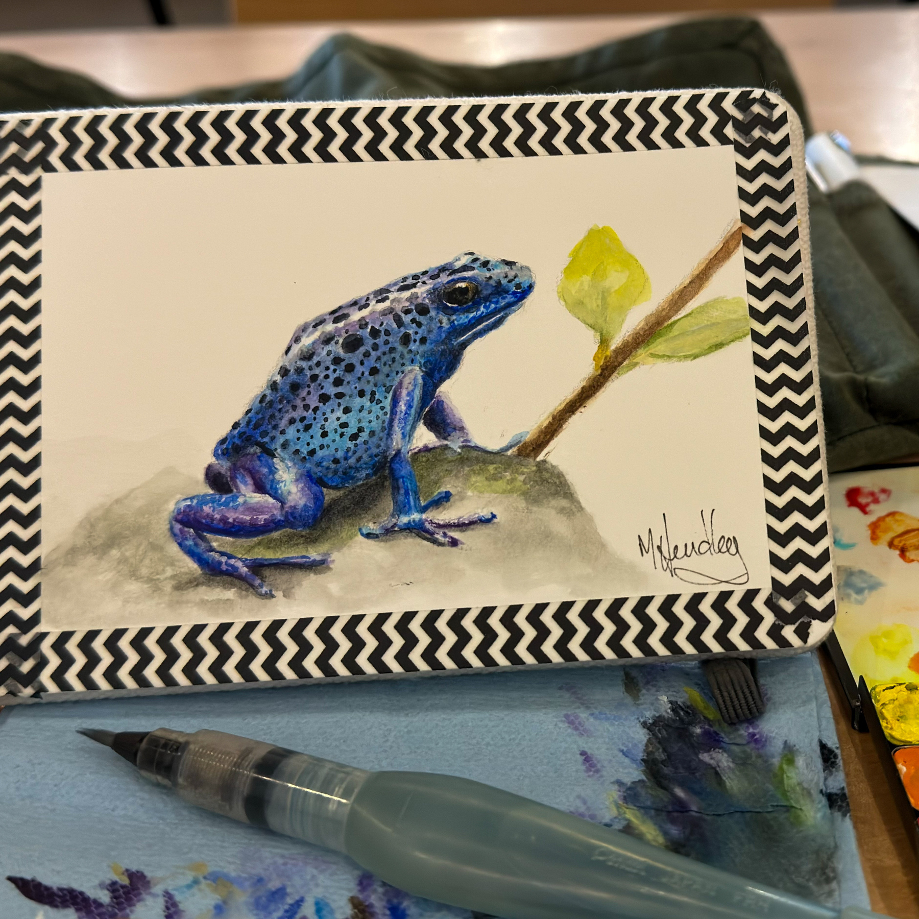 Watercolor painting of a blue poison dart frog perched on a rock with a single yellow-green leaf in the background, presented on a sketchbook with a chevron pattern border. Art supplies, including a water brush pen, are visible around the sketchbook.
