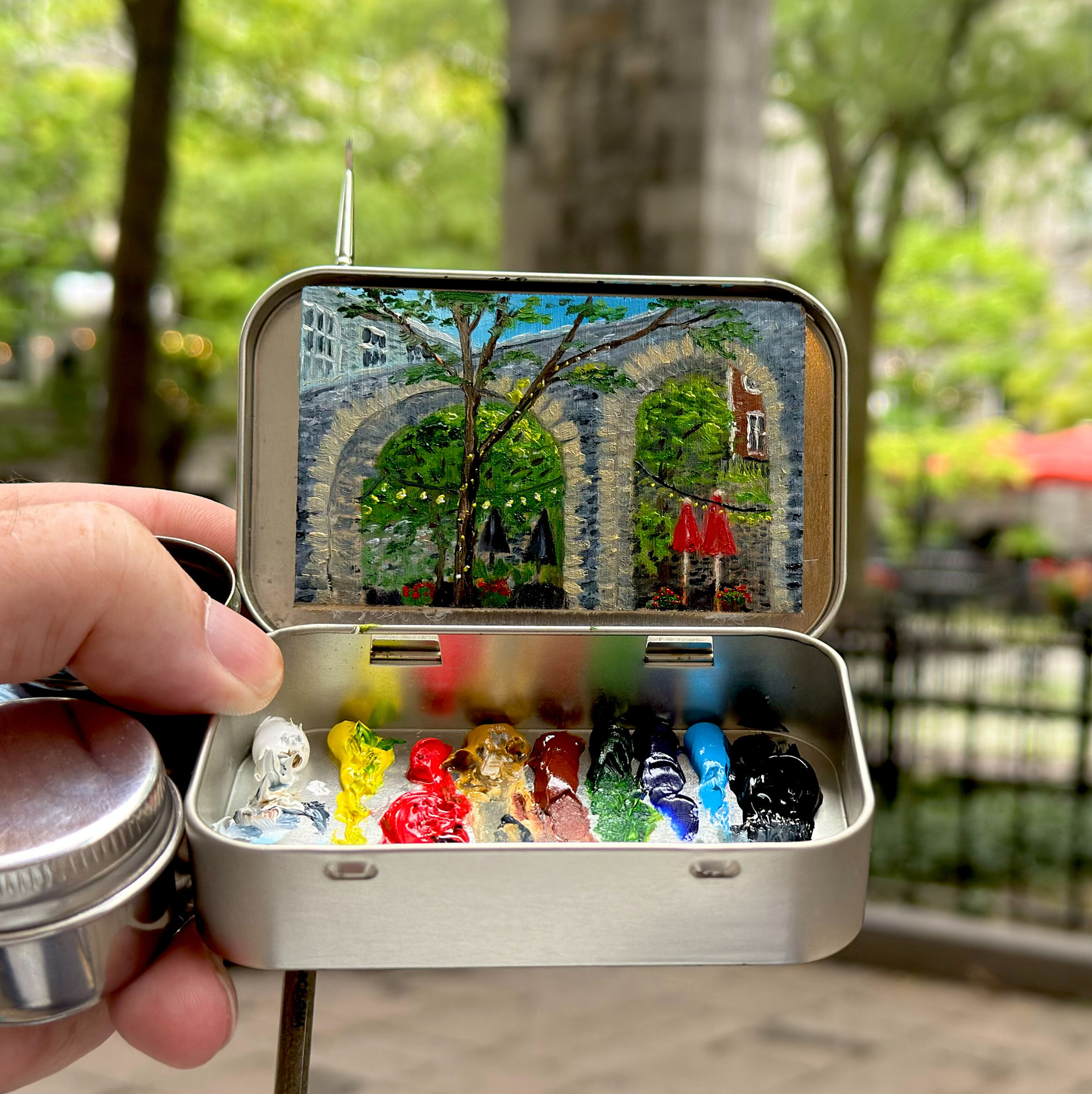 A miniature painting of a park scene is displayed inside a small open tin case with various colors of paint.