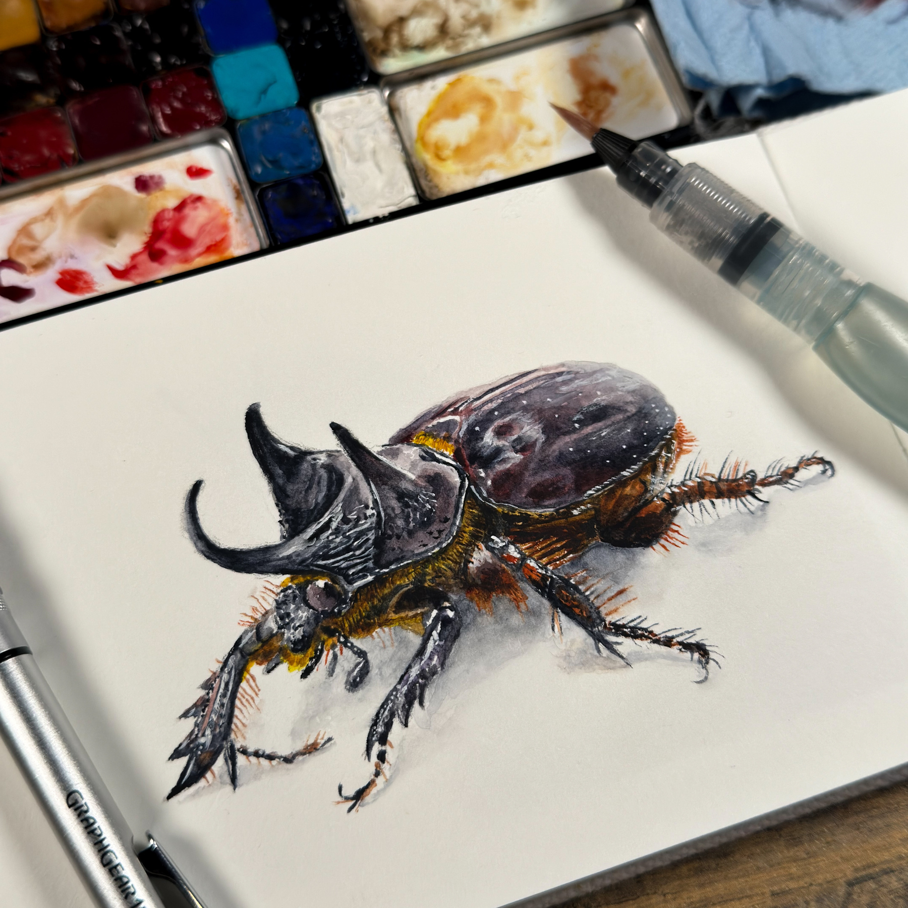 Watercolor illustration of a detailed box beetle on a white sketchbook page, accompanied by a painter's palette with a variety of colors, a water brush pen, and a pencil.