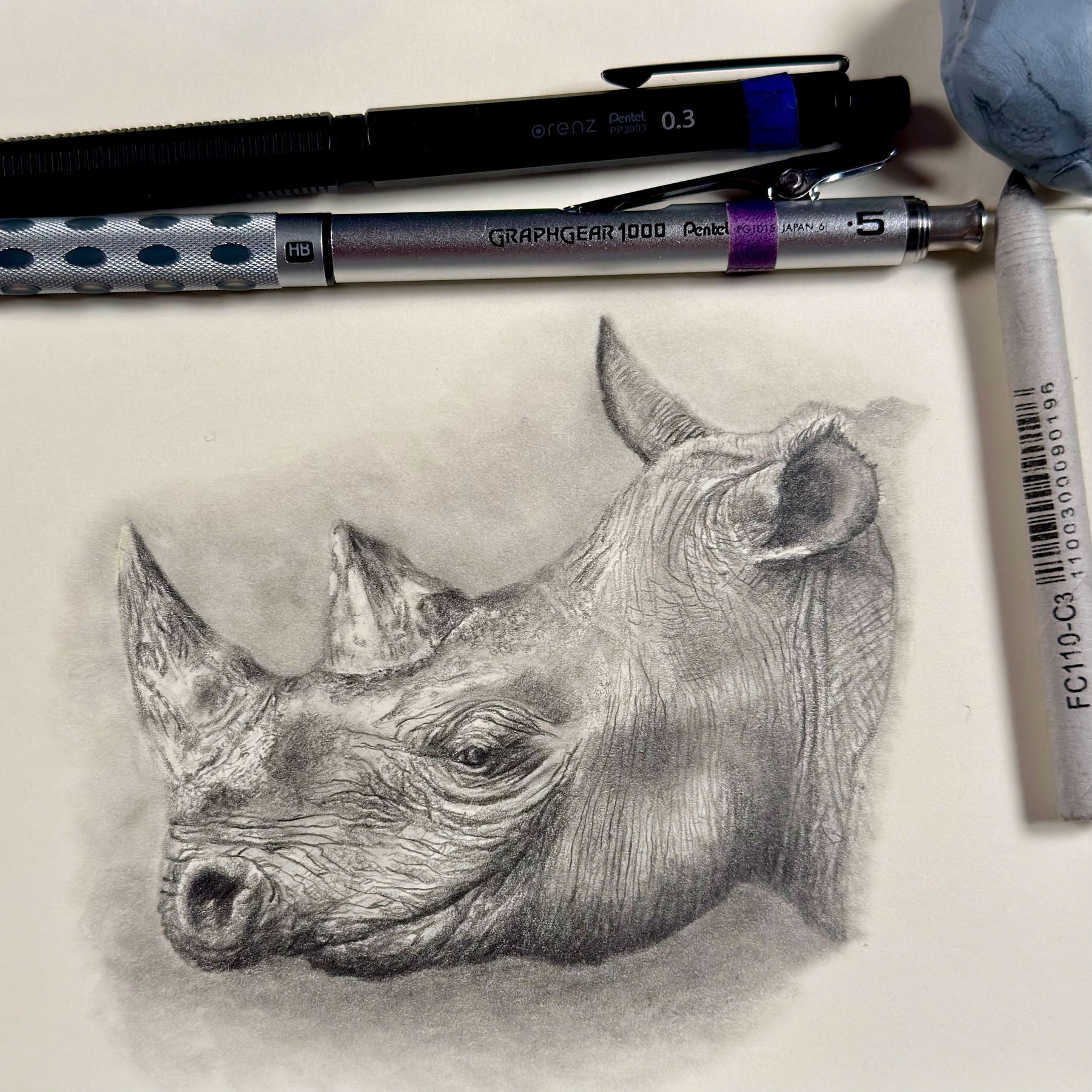 A detailed graphite sketch of a rhinoceros head, capturing the texture of its skin and the shape of its horn. Nearby, two Pentel drawing tools—a mechanical pencil, a drafting pencil, and a kneaded eraser—are placed above the artwork.