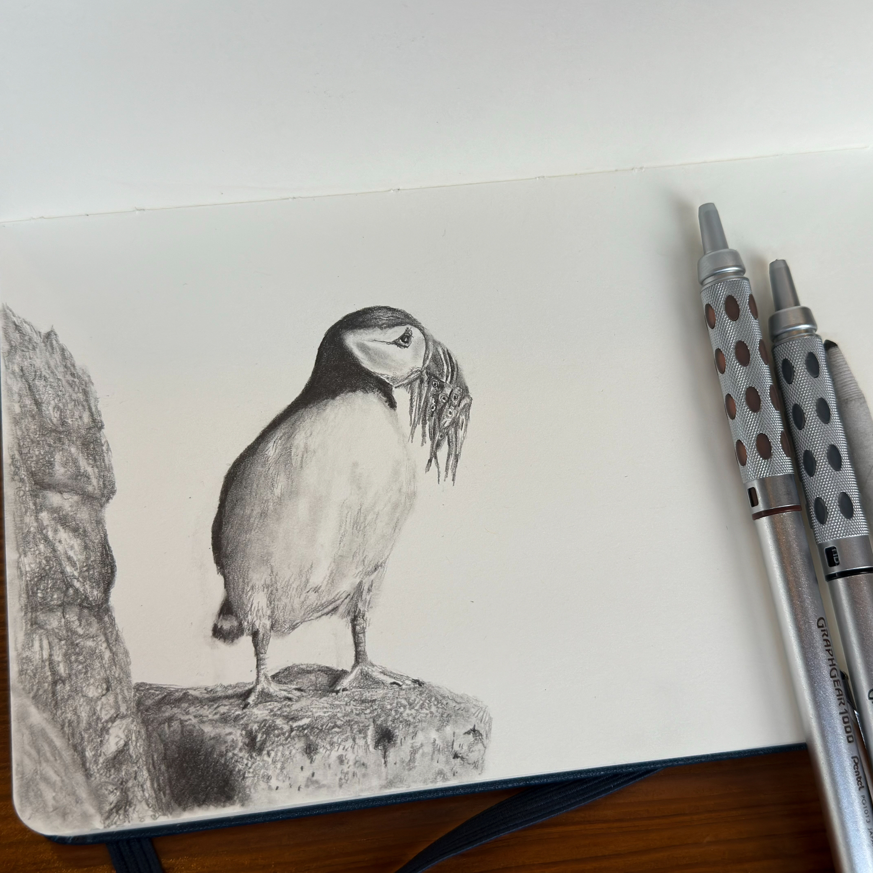 A pencil drawing of a puffin perched on a rock, with several fish in its beak, showcased on an open sketchbook next to two mechanical pencils on a wooden surface.