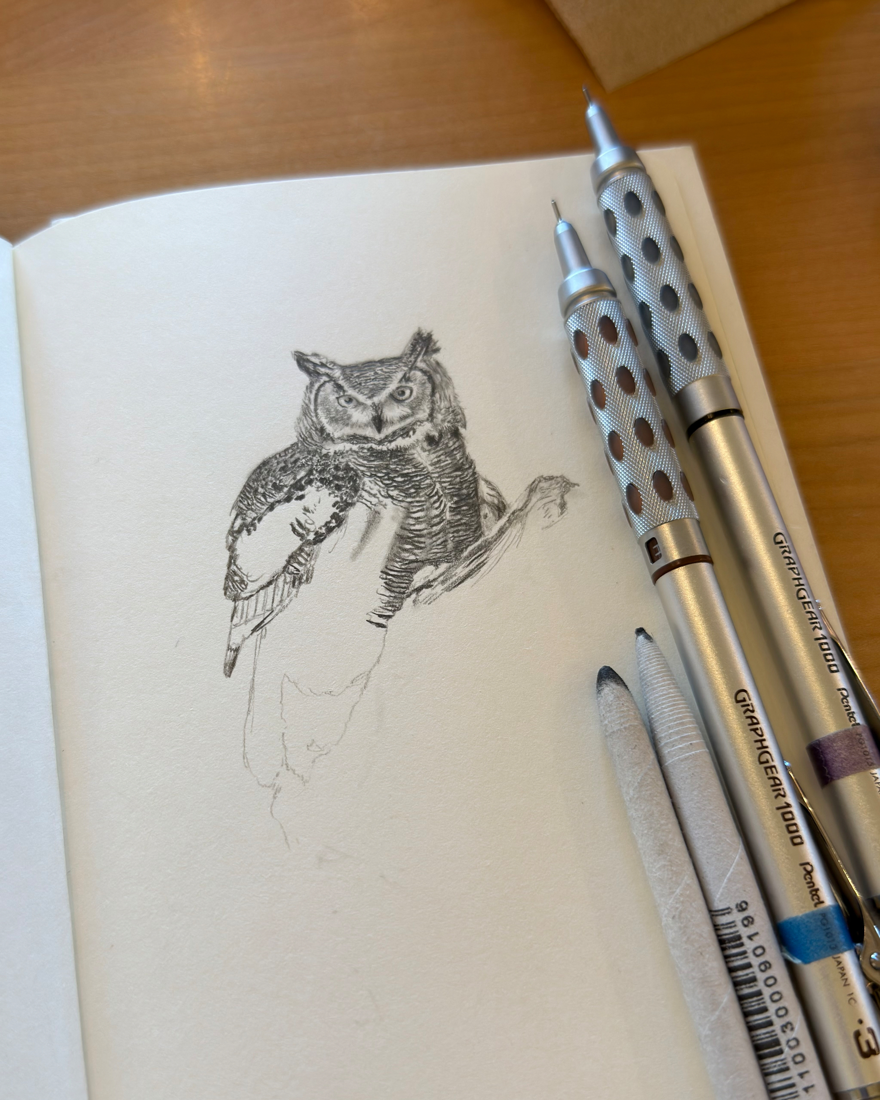 Open sketchbook on a wooden table with a partially finished graphite drawing of a great horned owl; two mechanical pencils and blending stumps lie beside the page.