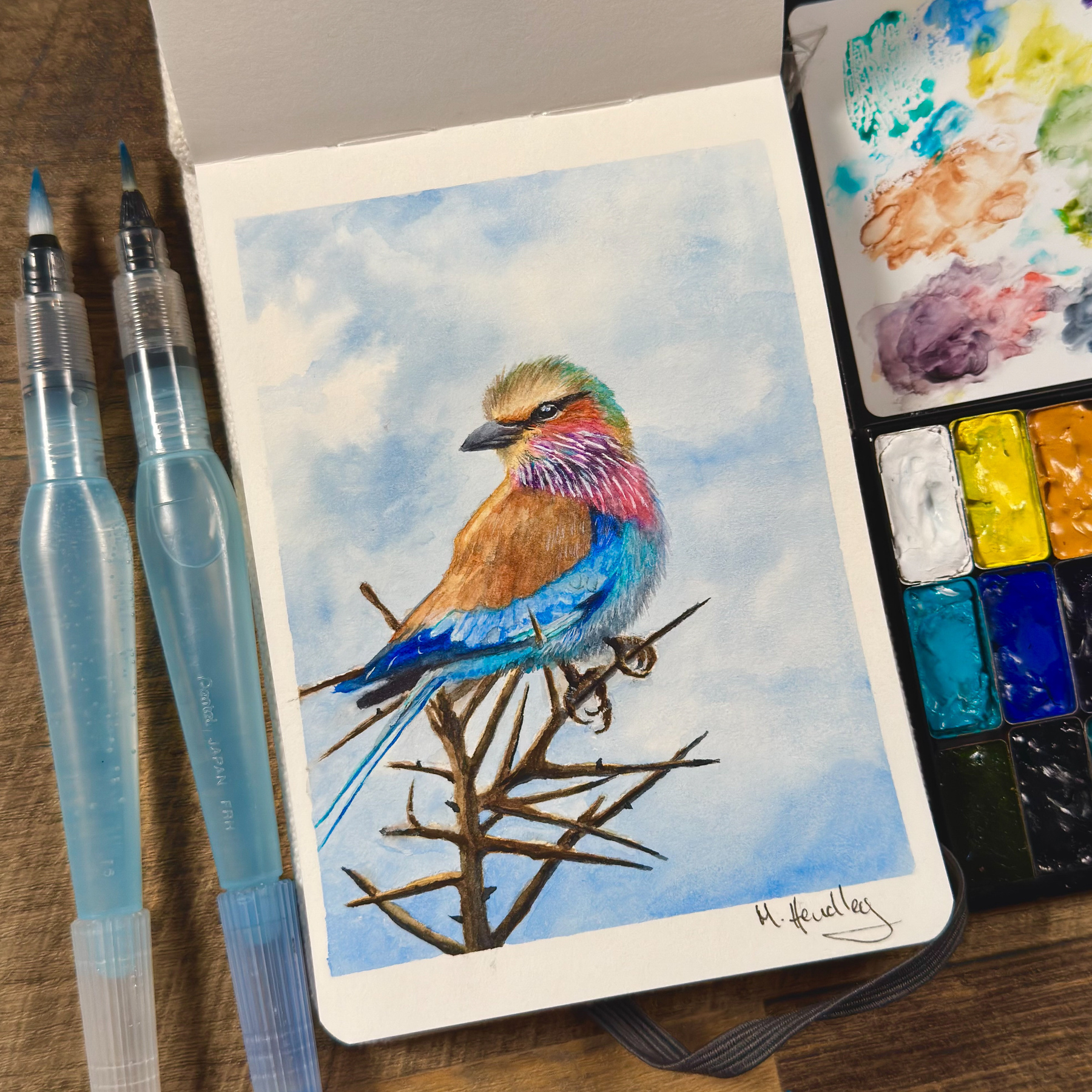 A watercolor painting of a vibrant lilac-breasted roller perched on a thorny branch, displayed in an open sketchbook. The bird has vivid blue, purple, pink, and brown tones, with a soft sky in the background. Nearby are water brushes and a watercolor palette.&10;