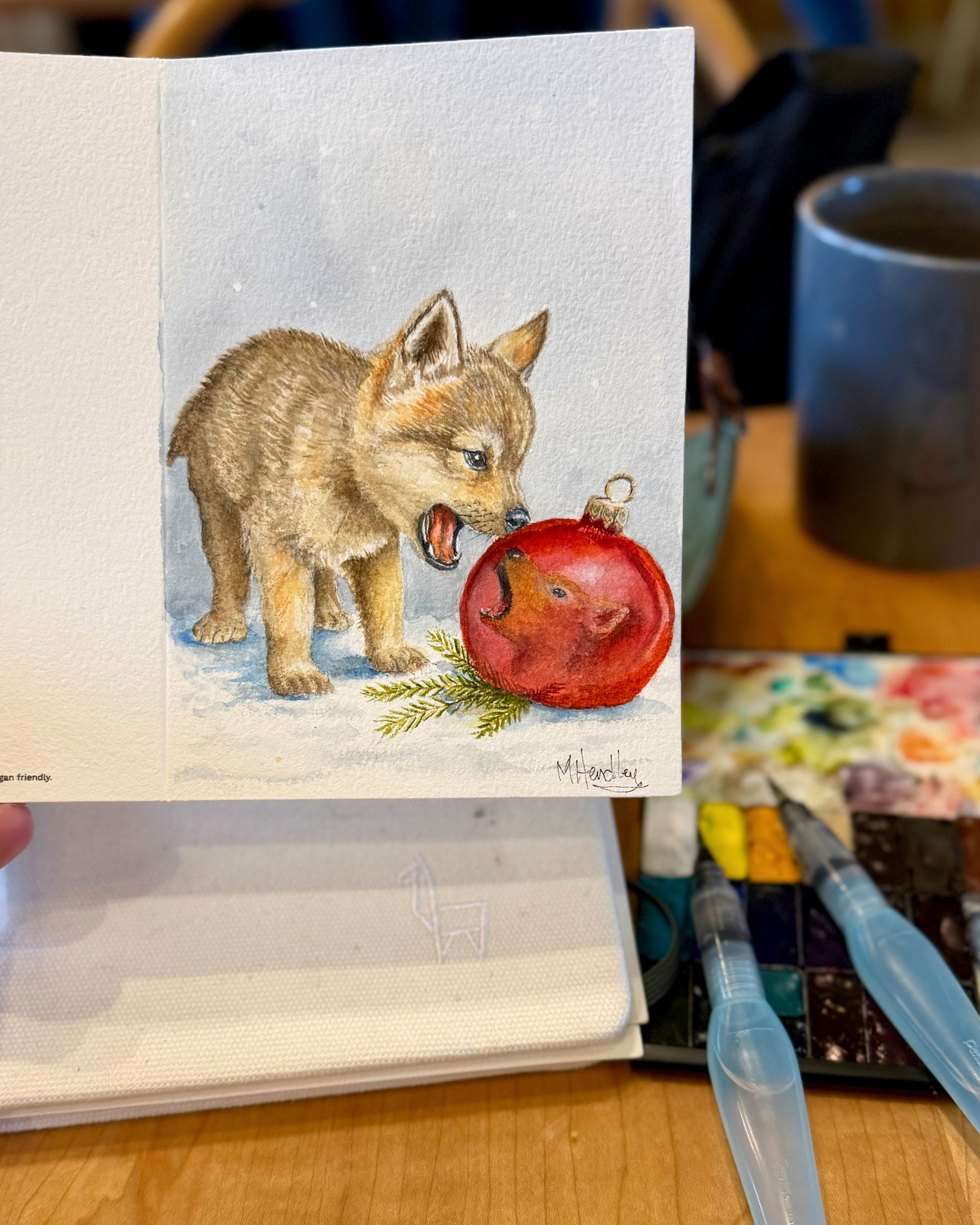 A watercolor painting of a curious wolf pup inspecting a shiny red ornament that reflects its own face. The scene is set against soft snow with a pine sprig nearby. The painting is shown on a handmade greeting card.&10;
