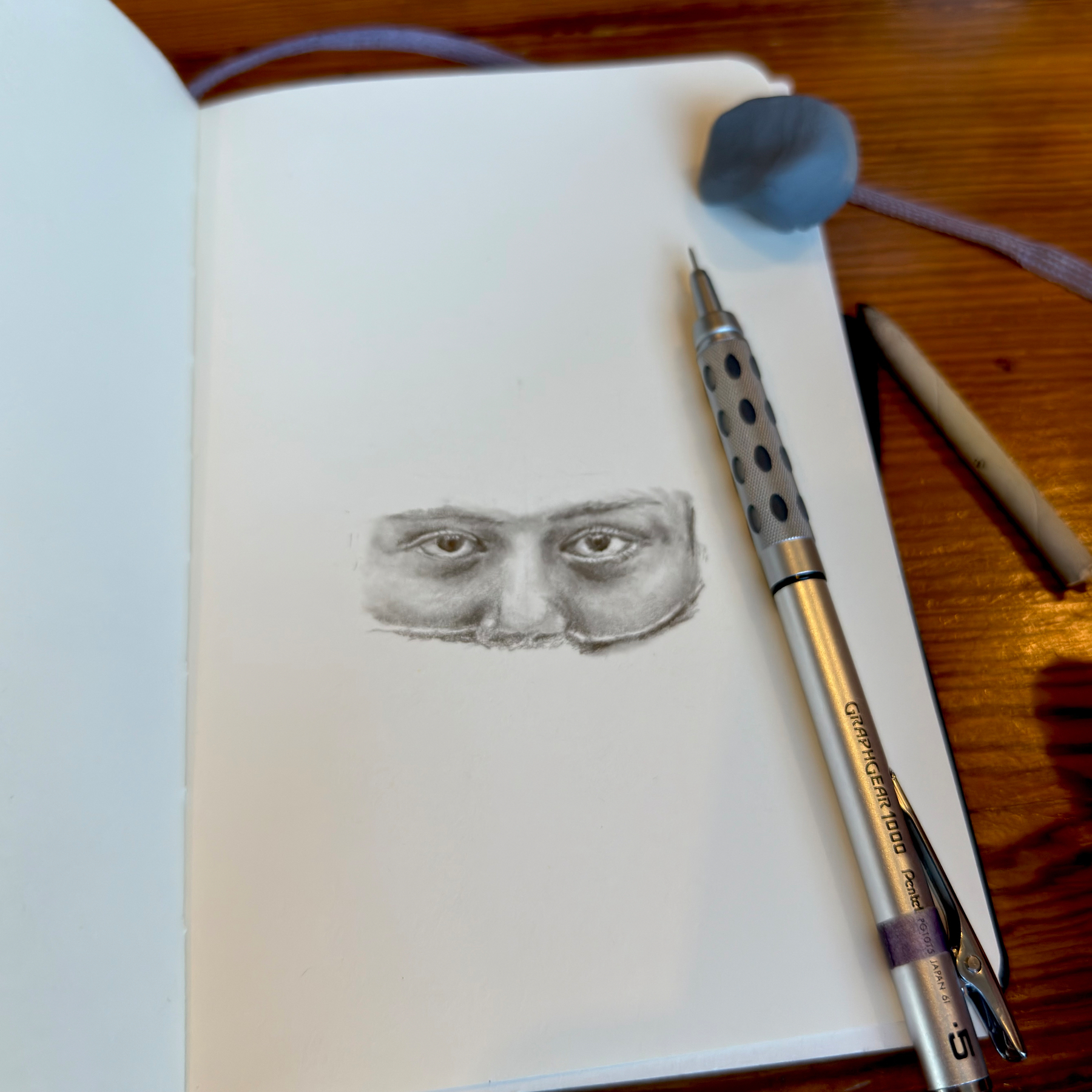 A graphite drawing in progress in a sketchbook shows a pair of realistic human eyes and the bridge of a nose emerging from the page. A mechanical pencil with a metal grip, a kneaded eraser, and a blending stump rest nearby on a wooden table, suggesting an artist’s workspace in use.