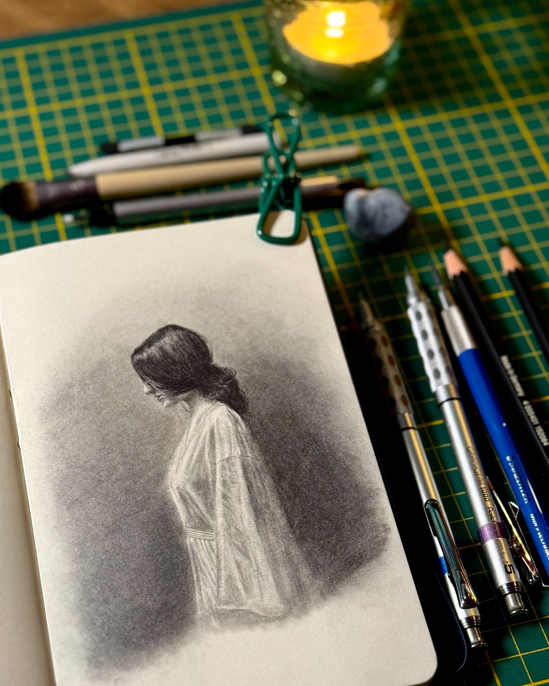 A graphite sketch of a woman in profile with her head slightly bowed, wearing a loose garment. The shading is soft, with smooth gradients blending into the paper’s texture. Surrounding the sketchbook are Graphgear pencils, clutch pencils, Faber-Castell Pitt graphite, graphite powder, brushes, and an eraser.