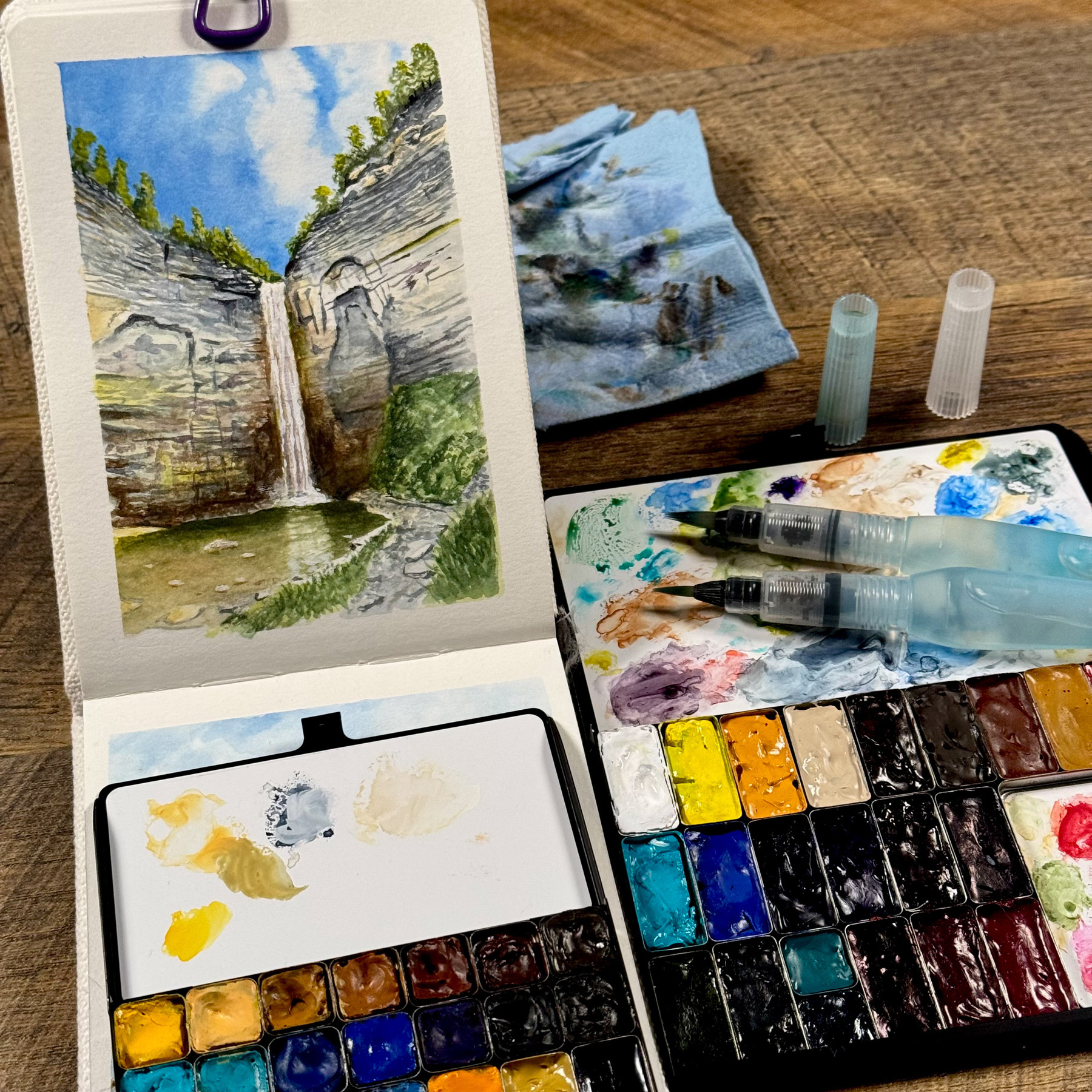 A vibrant watercolor sketch of Taughannock Falls in a sketchbook, surrounded by a compact watercolor palette, water brushes, and a blue-stained paper towel. The waterfall cascades between towering rock cliffs, framed by trees and a bright blue sky.