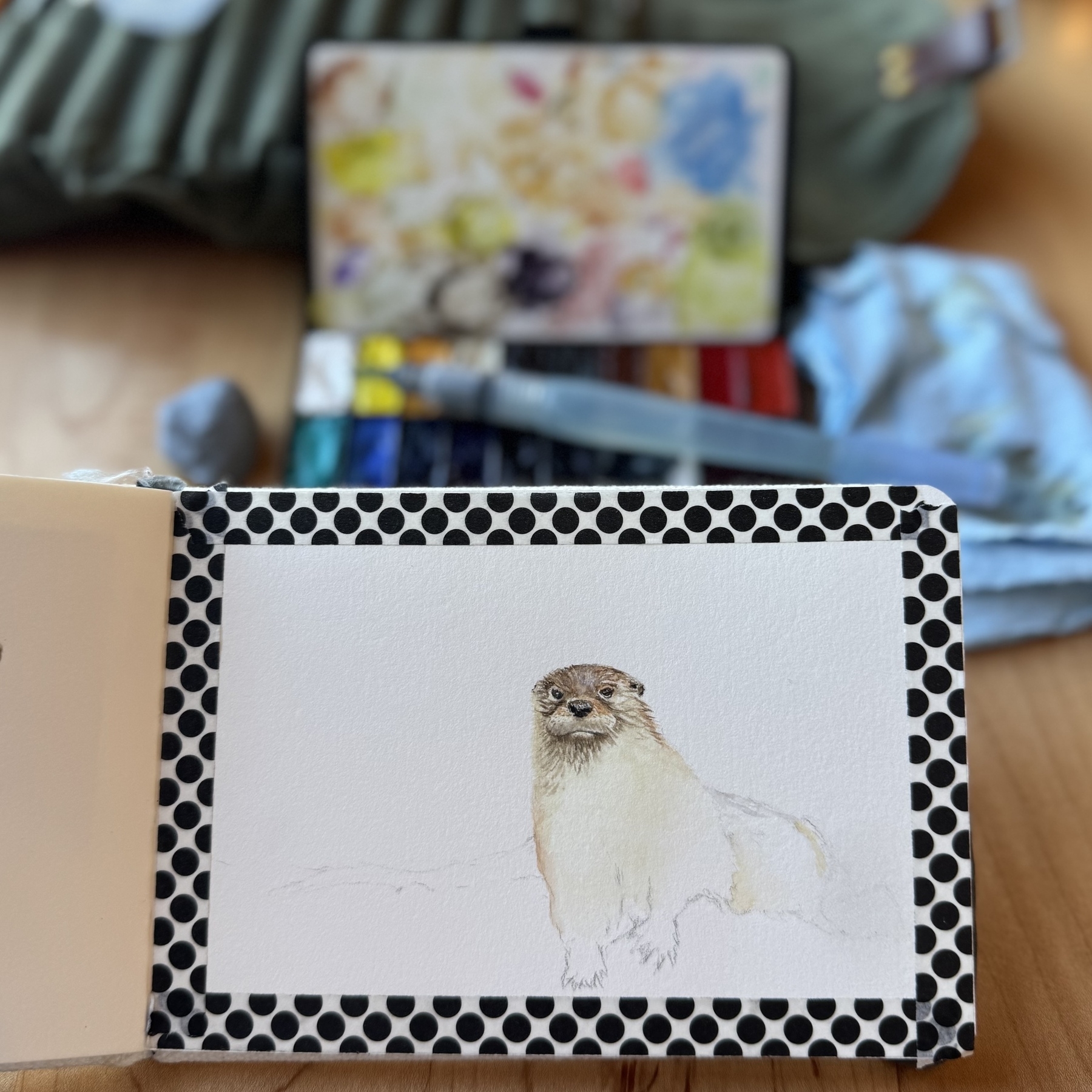 A watercolor sketch of an otter in progress in a sketchbook. Palette and water brush in the background. 