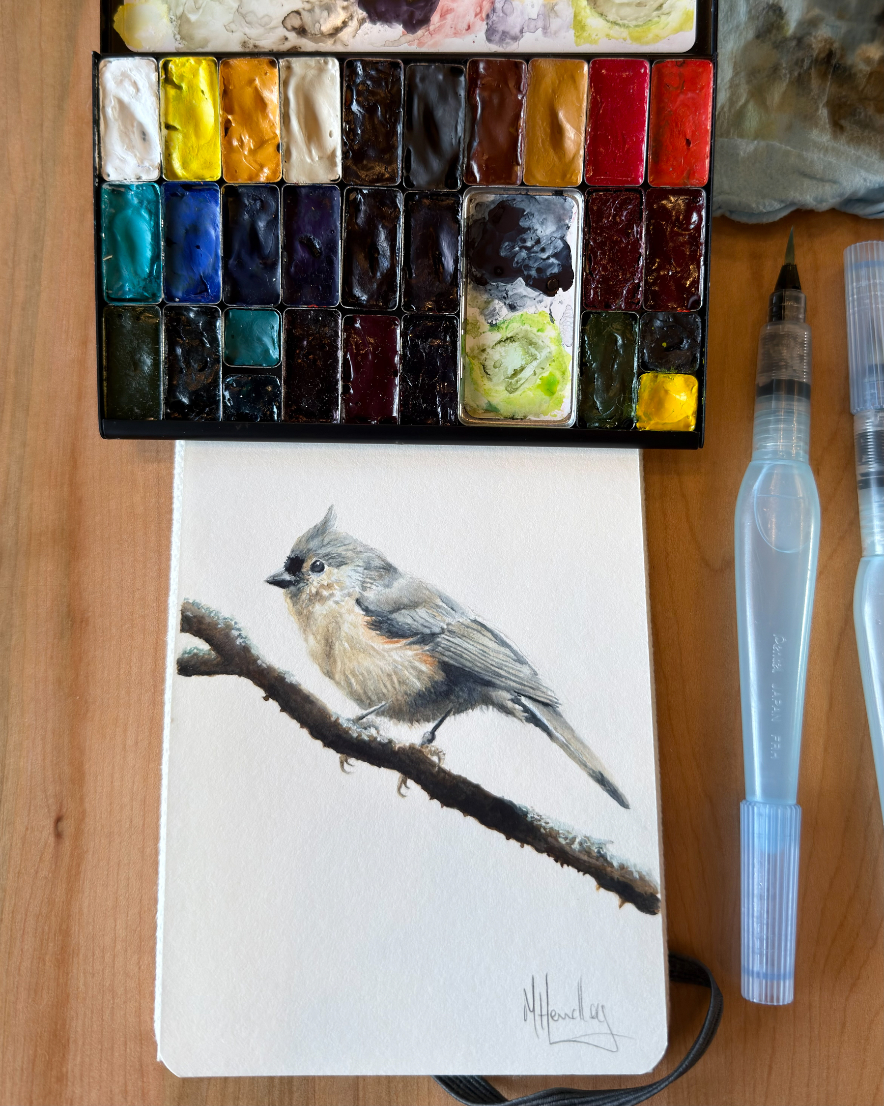 Watercolor painting of a tufted titmouse perched on a branch, painted in a sketchbook with visible palette and water brushes beside it.
