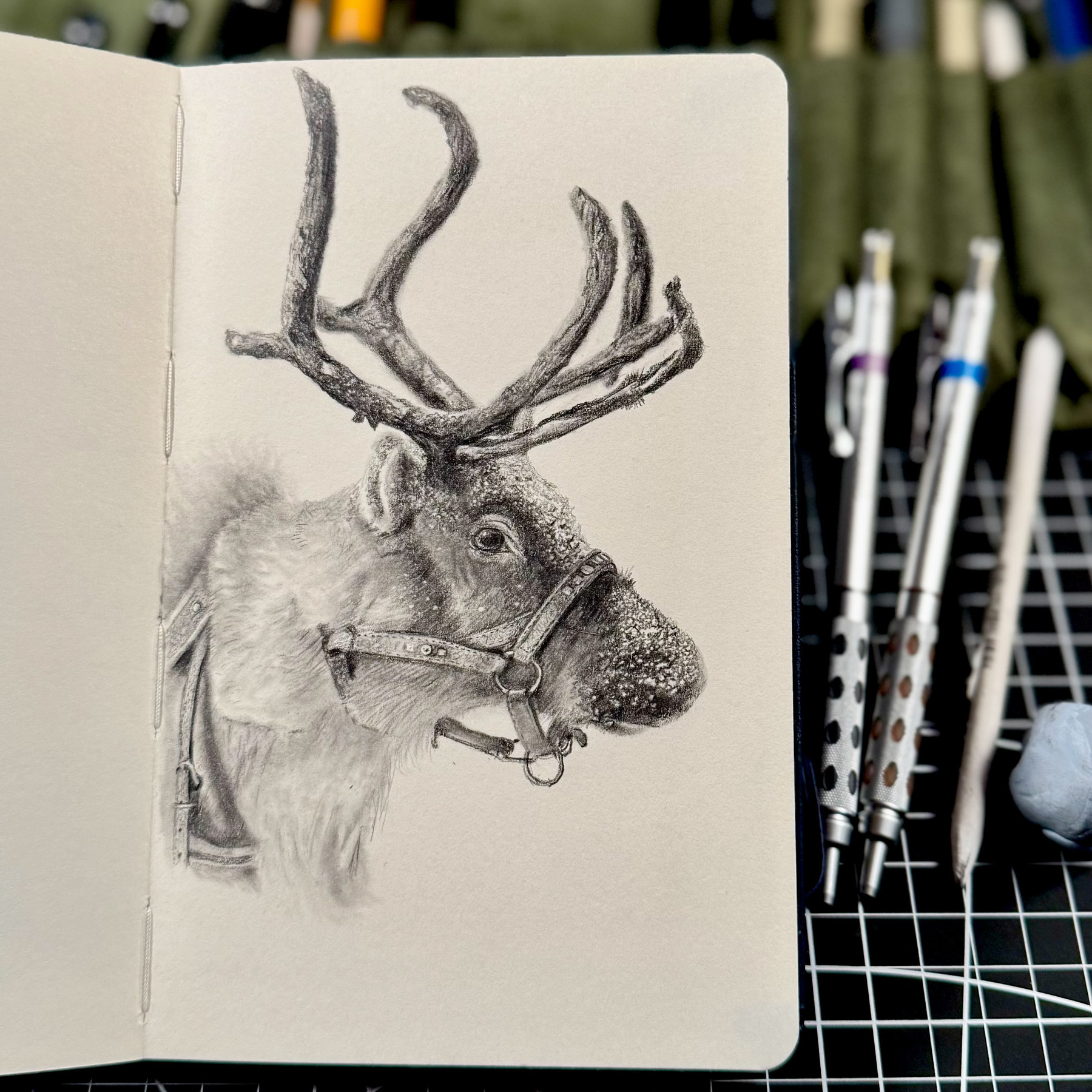 A realistic graphite drawing of a reindeer in a sketchbook, capturing intricate details of its fur, antlers, and a leather halter. Snowflakes are visible on its muzzle. The sketchbook is open on a gridded surface, surrounded by mechanical pencils and blending tools.