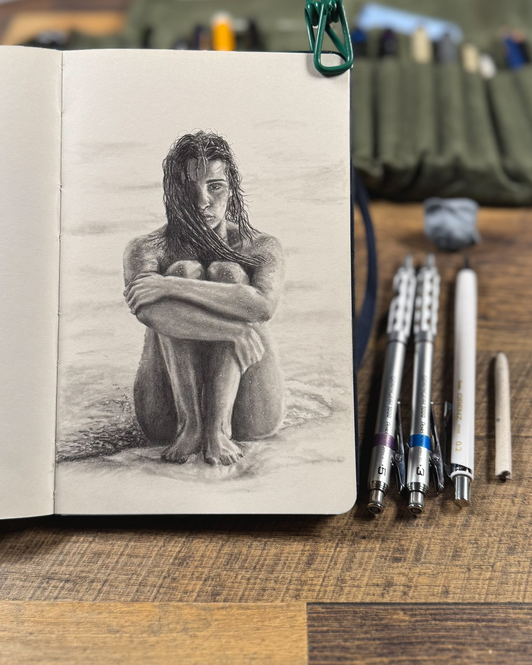 A graphite pencil drawing of a woman sitting on a beach with wet hair, arms wrapped around her knees. Her gaze is intense, and sand clings to her skin. The drawing captures light and shadow masterfully, with a blurred ocean in the background. Art tools are laid out beside the sketchbook.