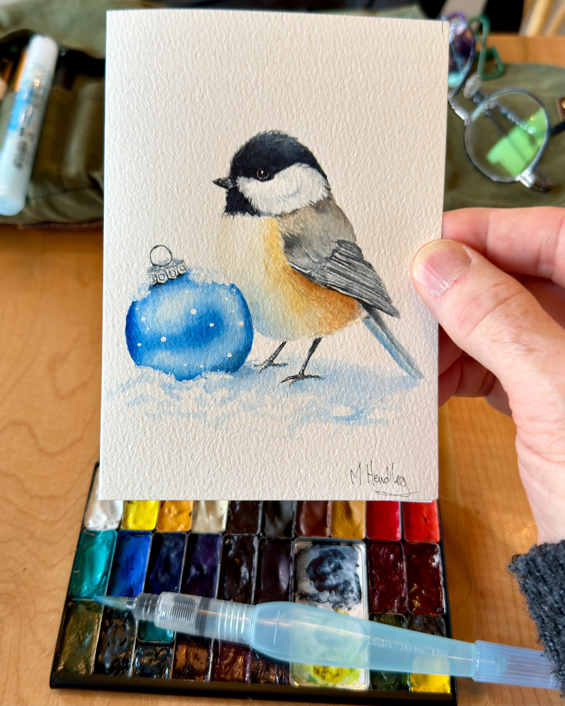 Watercolor painting of a chickadee standing in snow next to a blue ornament, with soft shading and fine details, held above a watercolor palette.