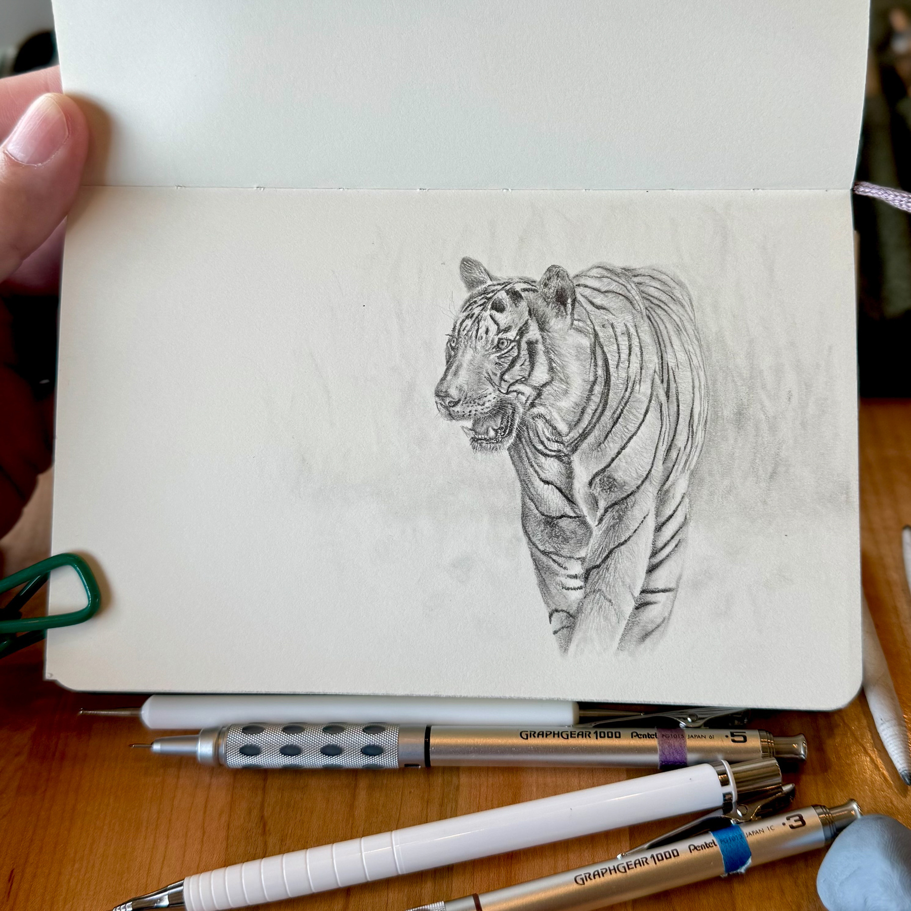 Realistic graphite sketch of a Bengal tiger mid-step, mouth slightly open in a soft snarl. Fine pencil shading captures its stripes and muscular form. Drawn in a sketchbook with mechanical pencils visible on the table.