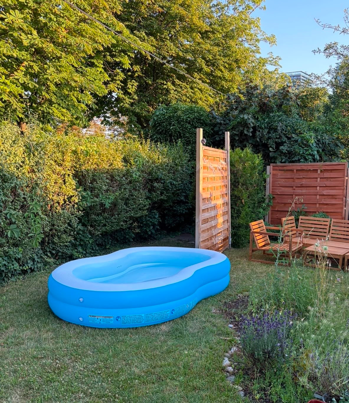 A small, blue inflatable pool is set up on a grassy lawn next to wooden outdoor furniture and surrounded by lush greenery.