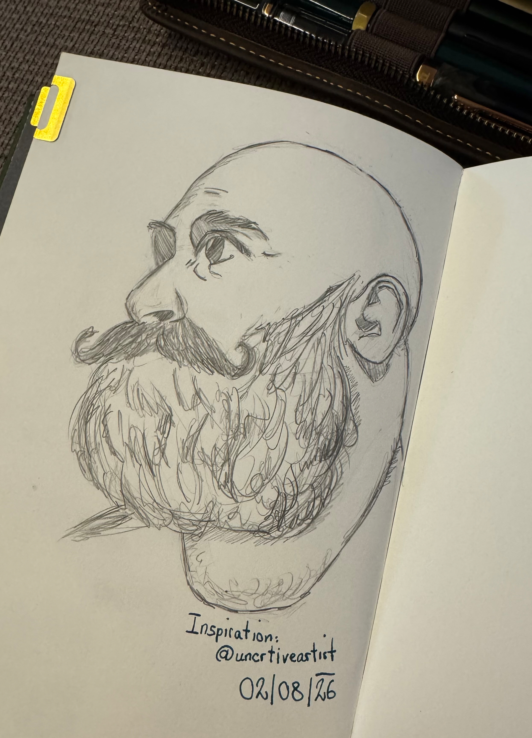 A pencil sketch in a notebook shows a side profile of a bald man with a full beard and mustache, accompanied by a note crediting inspiration to @uncrtiveartist and dated 02/08/26.