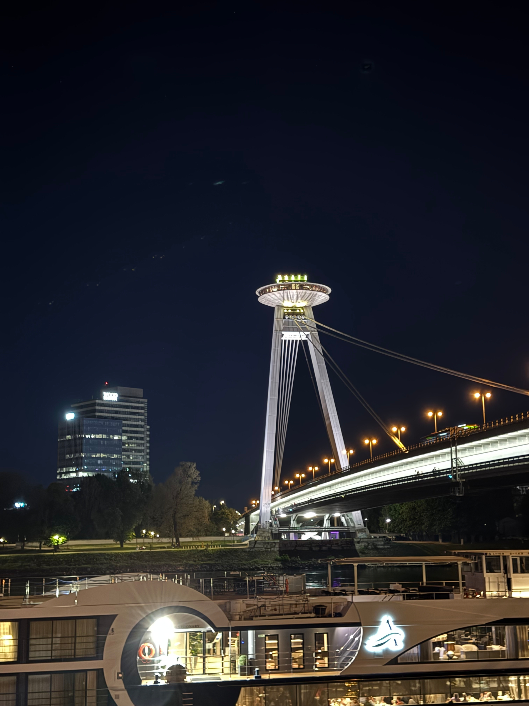 A modern cable-stayed bridge with a UFO-shaped observation tower, illuminated at night, stretches across a river with a nearby lit building and trees.