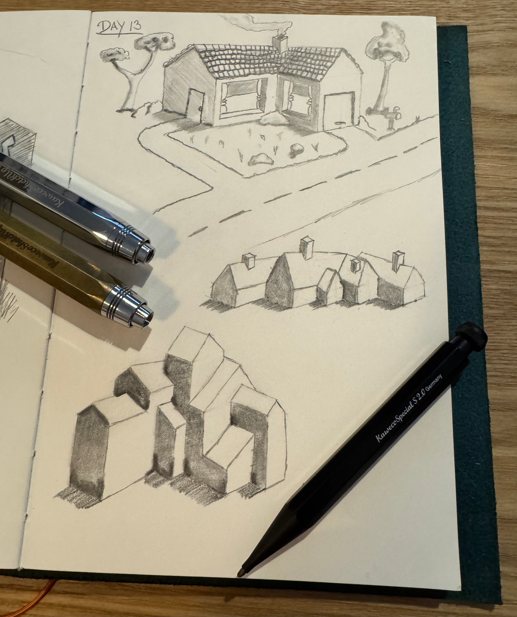A sketchbook page displays pencil drawings of houses, geometric shapes, and includes two pens and a pencil on top.