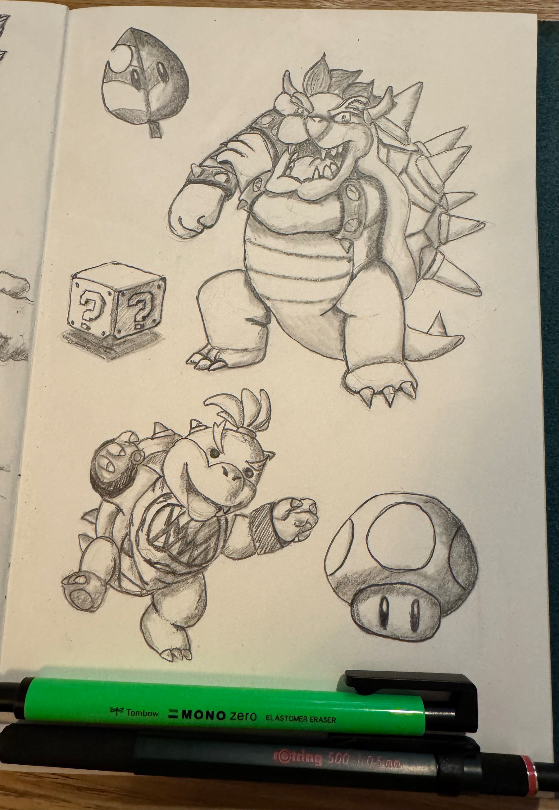 A sketchbook page features pencil drawings of cartoon-style characters and objects, including a spiked turtle-like creature, a block with a question mark, a helmet, and a mushroom, with pens resting on the page.