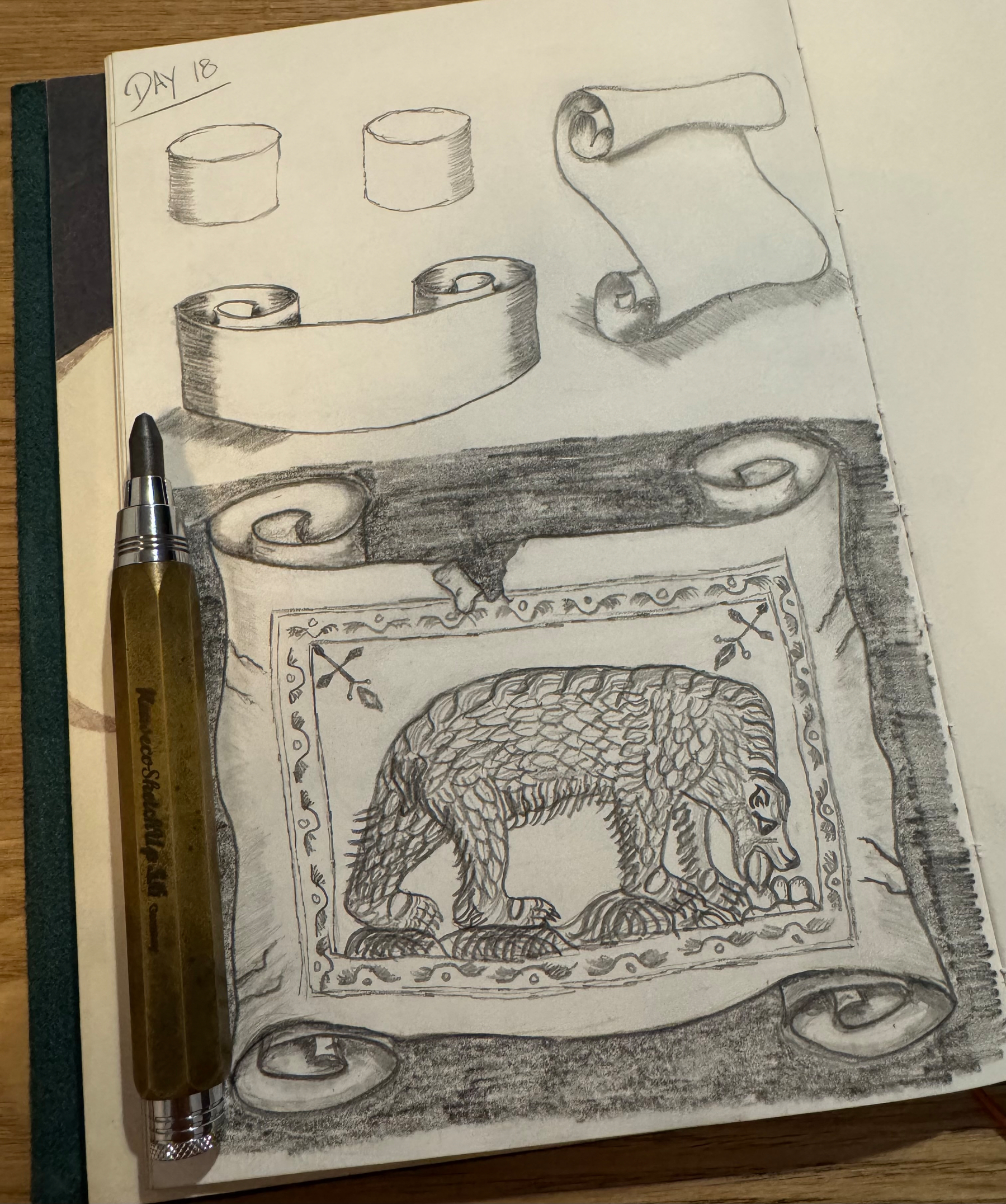 A sketchbook page features pencil drawings of cylindrical objects, a scroll, and a detailed rectangular rug with a bear design, alongside a pen resting on the paper.