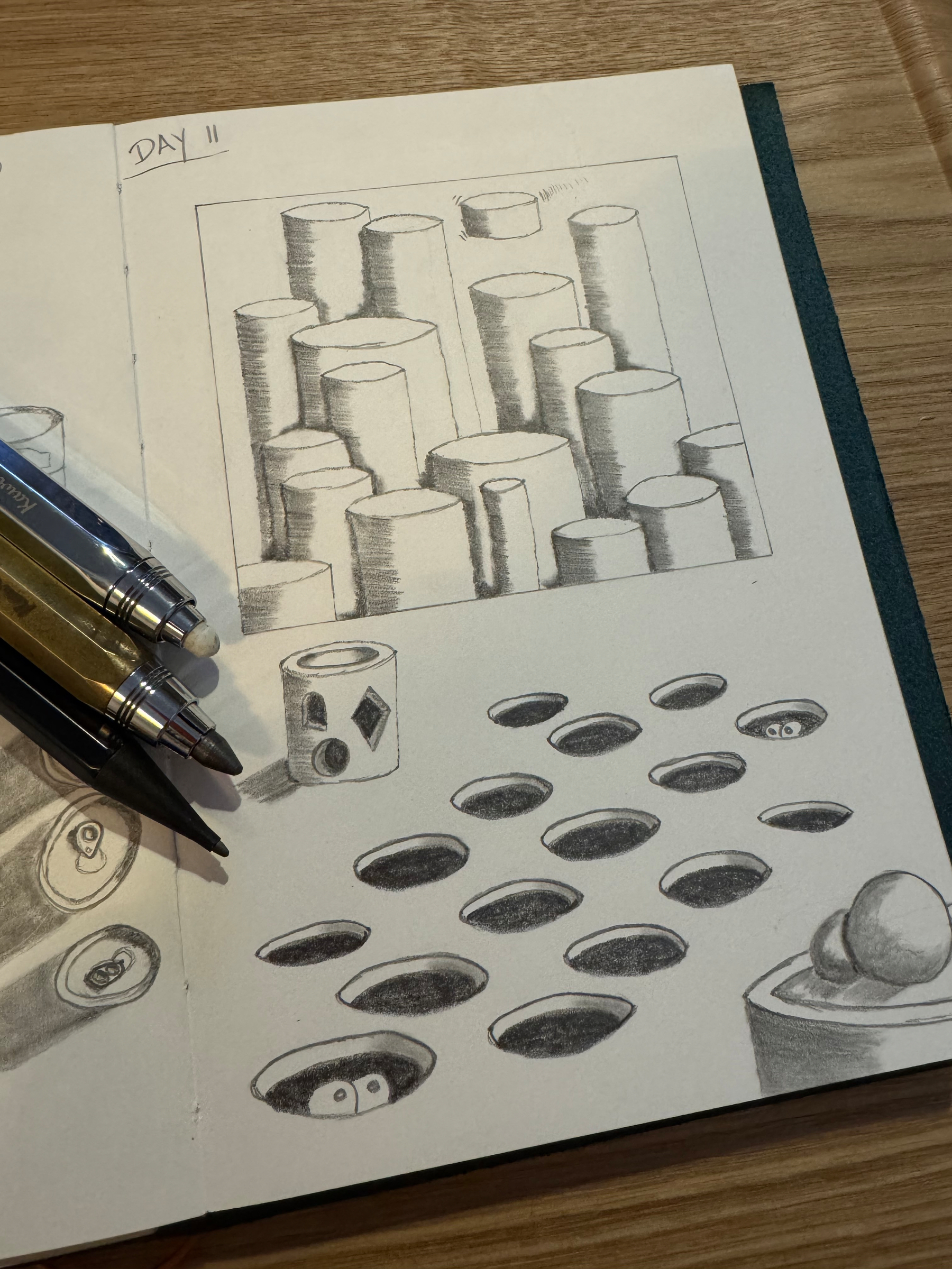 A sketchbook page features a drawing of cylindrical shapes, another of holes with eyes inside, and two pens placed on it.