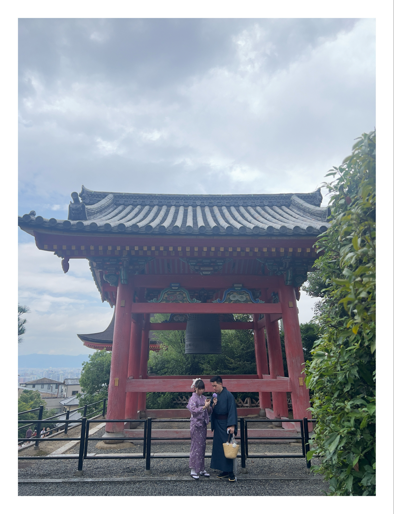 A man and a woman in traditional Japanese attire stand in front of a large red bell tower, with cloudy skies overhead and greenery to the right.