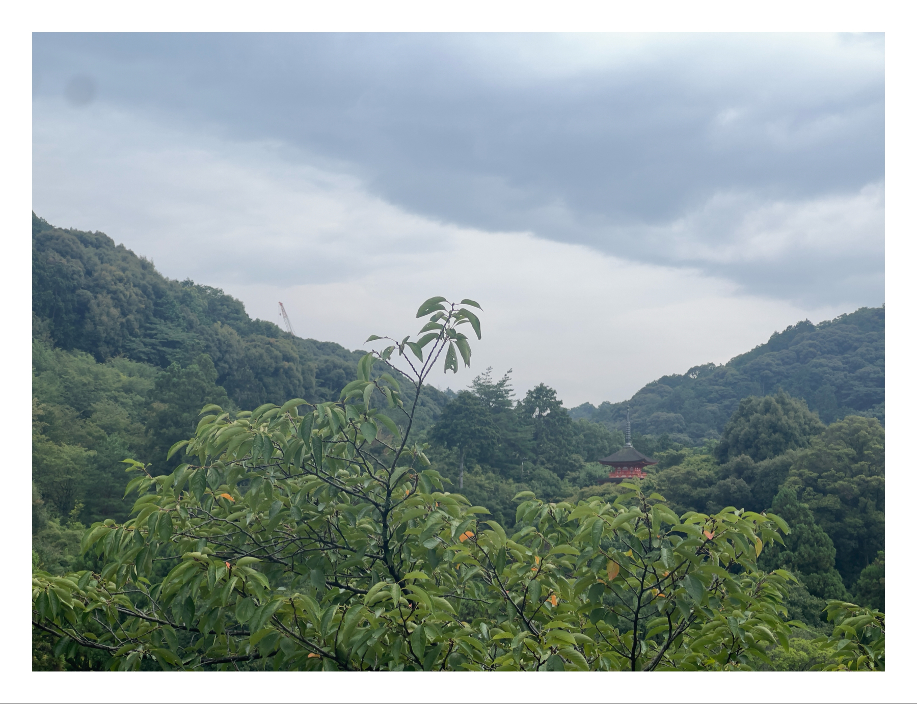 A serene landscape of lush green hills with a traditional Japanese pagoda nestled among the trees under a cloudy sky. A crane is visible in the distance on the left side, suggesting construction or maintenance activity.