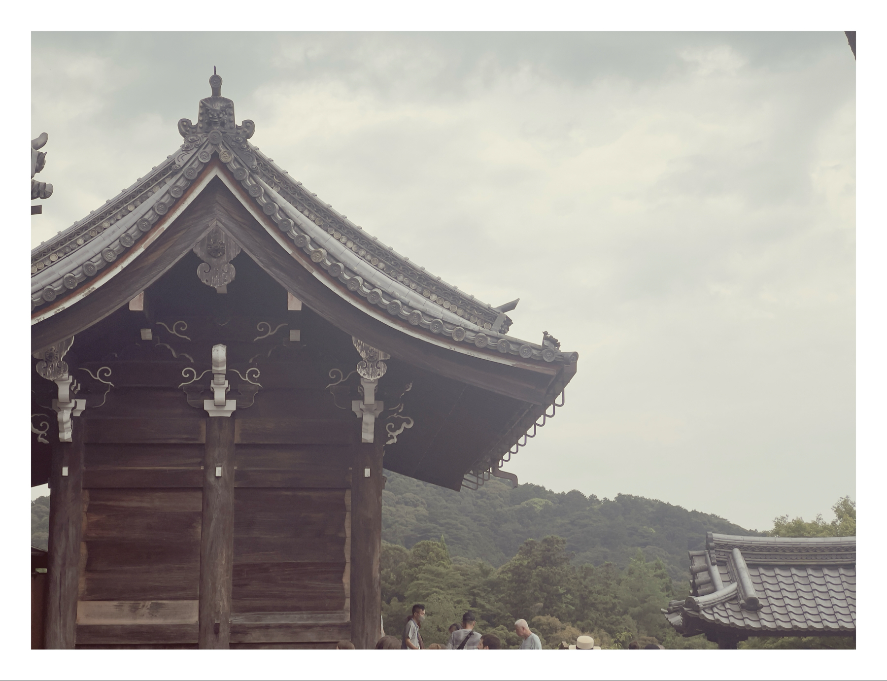 Traditional Japanese wooden structure with ornate roof and metal fixtures, set against a backdrop of lush green hills. A few people are visible at the bottom, suggesting a peaceful, historical setting.