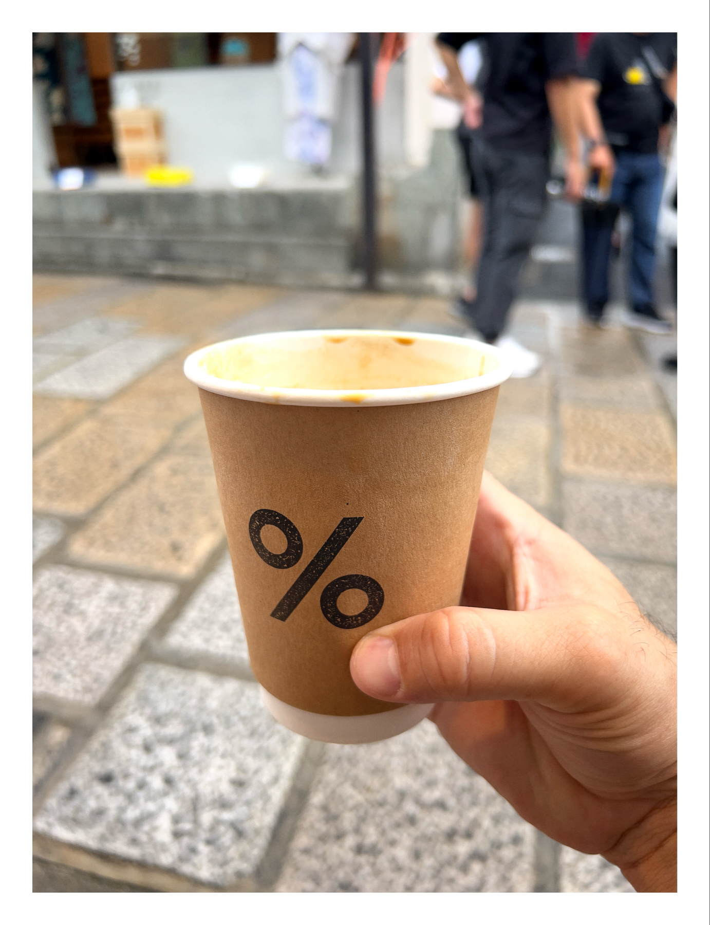 A hand holding a paper coffee cup with “%” logo printed on it, with an out-of-focus background featuring people walking on a street.