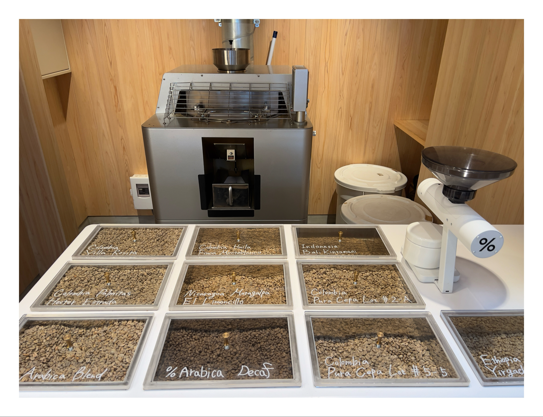 The image shows a self-service coffee bean selection area with a variety of coffee beans displayed in clear trays, each labeled with their origin and type. A coffee grinder is to the right, and a commercial coffee machine is at the back. 