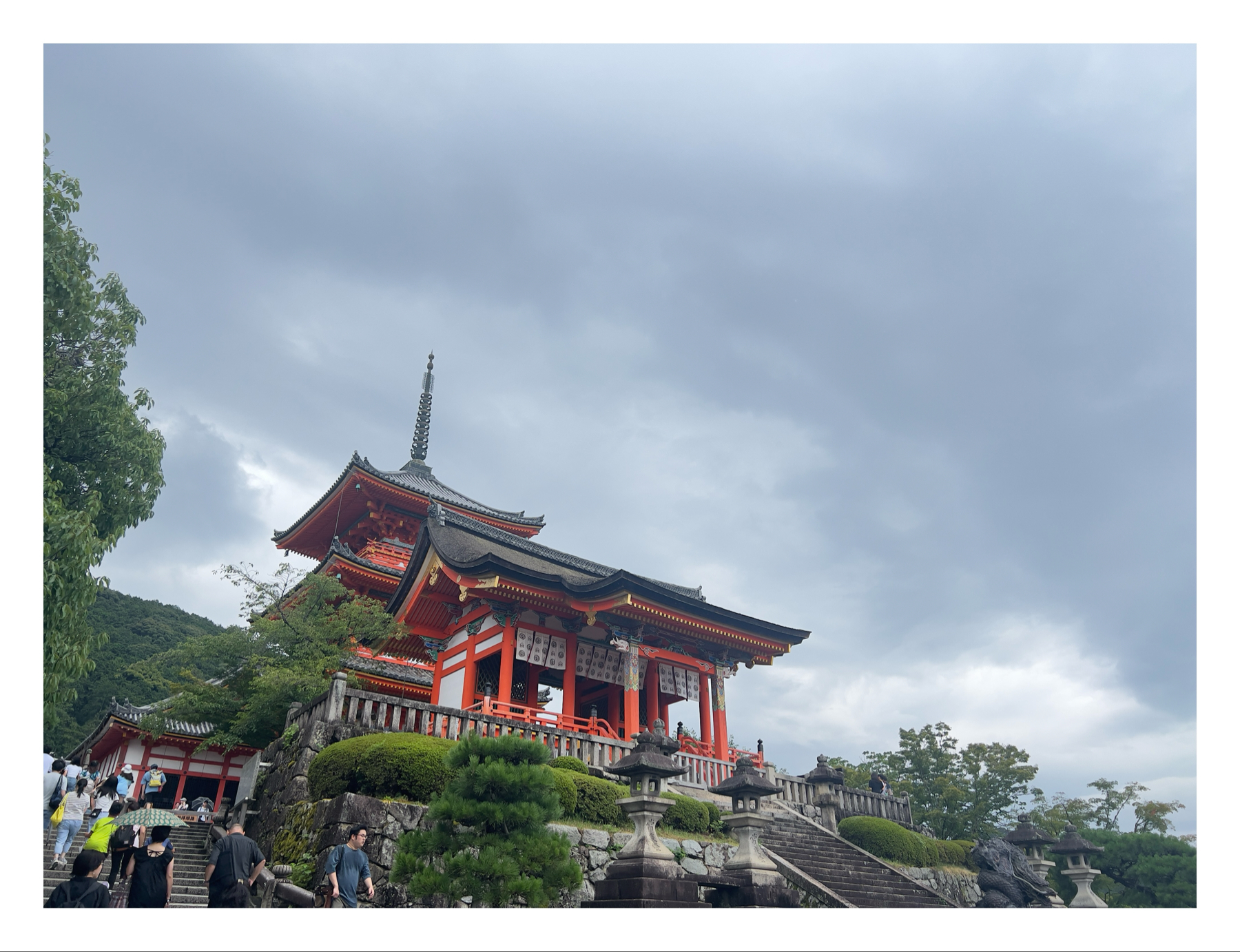 A traditional Japanese pagoda with bright red accents set against a cloudy sky, surrounded by lush greenery, with visitors ascending the stone steps.