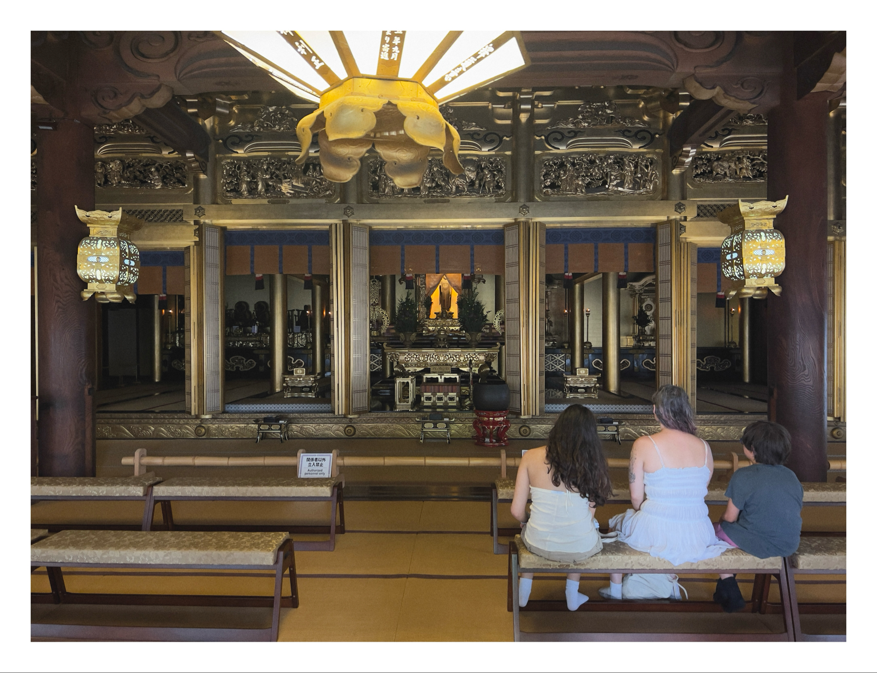 Three individuals seated on a bench in front of an ornate altar inside a temple, with intricate carvings and hanging lanterns visible.