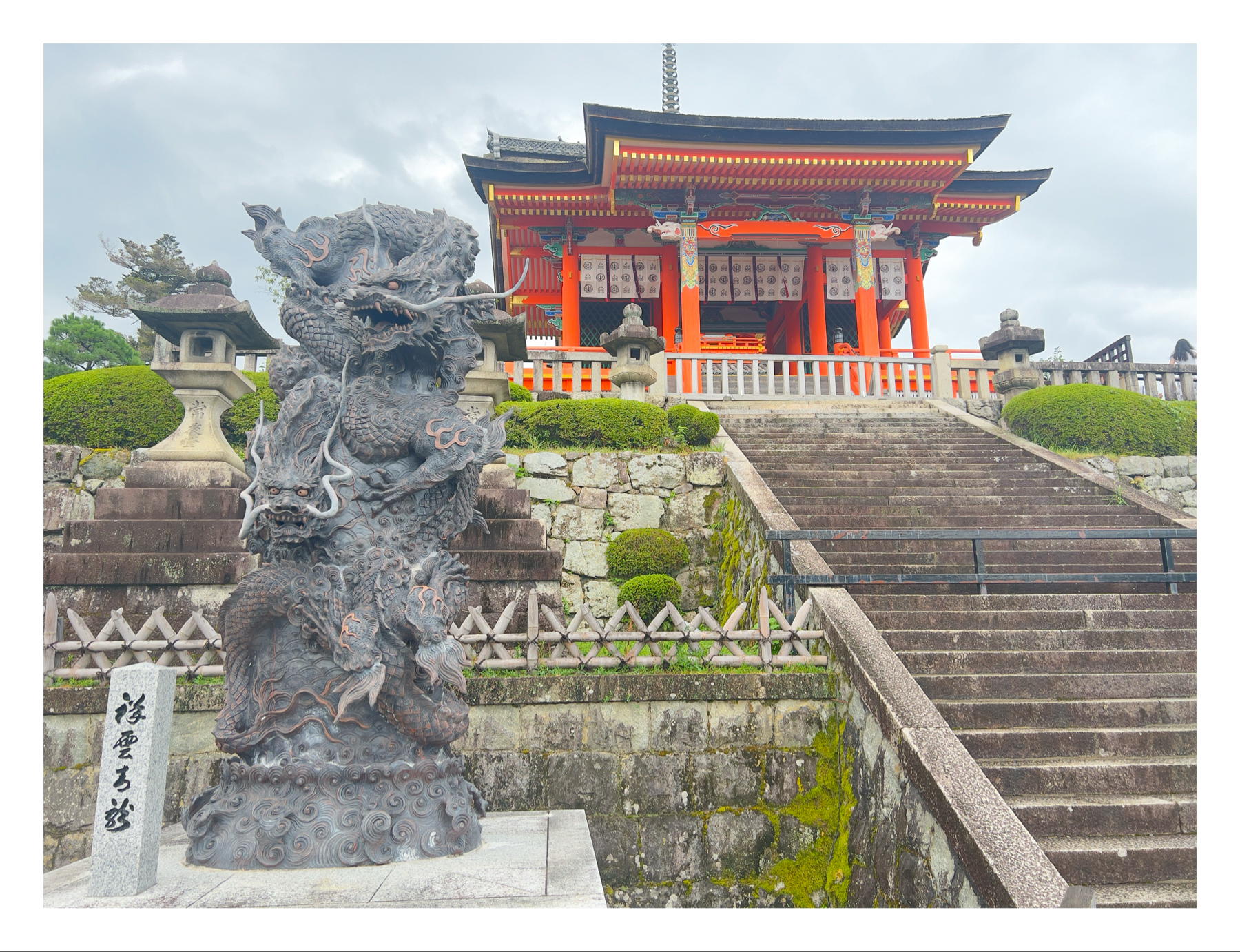 Sculpture of a dragon at the base of stone steps leading up to a traditional orange Japanese temple with decorative details and a cloudy sky in the background.