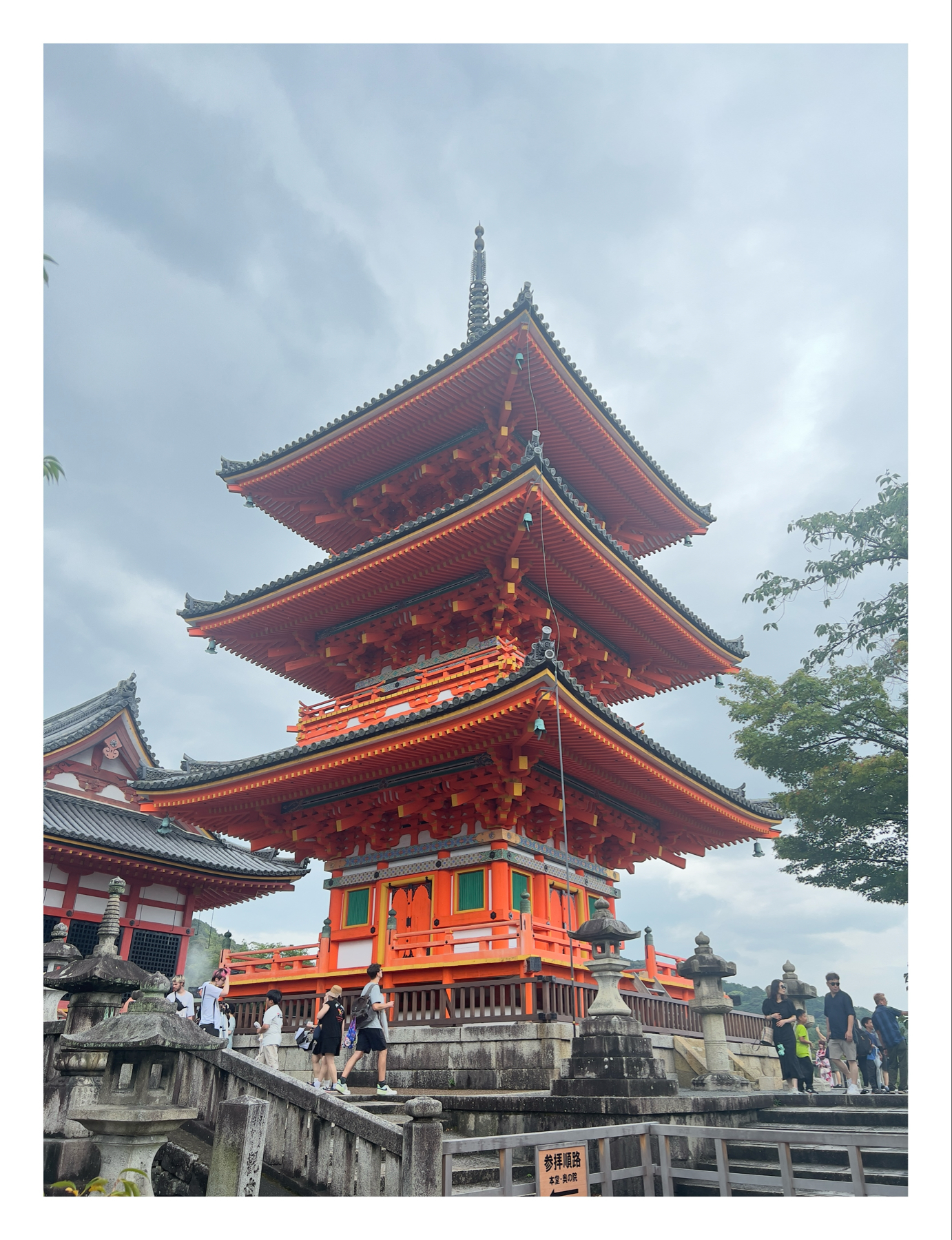 A vibrant orange, multi-tiered pagoda in a Japanese architectural style, with visitors walking and taking photos around the structure, set against a cloudy sky.