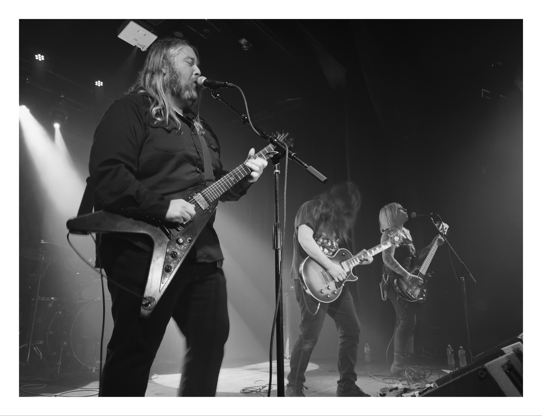 Black and white photo of three musicians performing on stage, with the central figure singing into a microphone and playing an electric guitar, flanked by two other guitarists engrossed in playing their instruments.