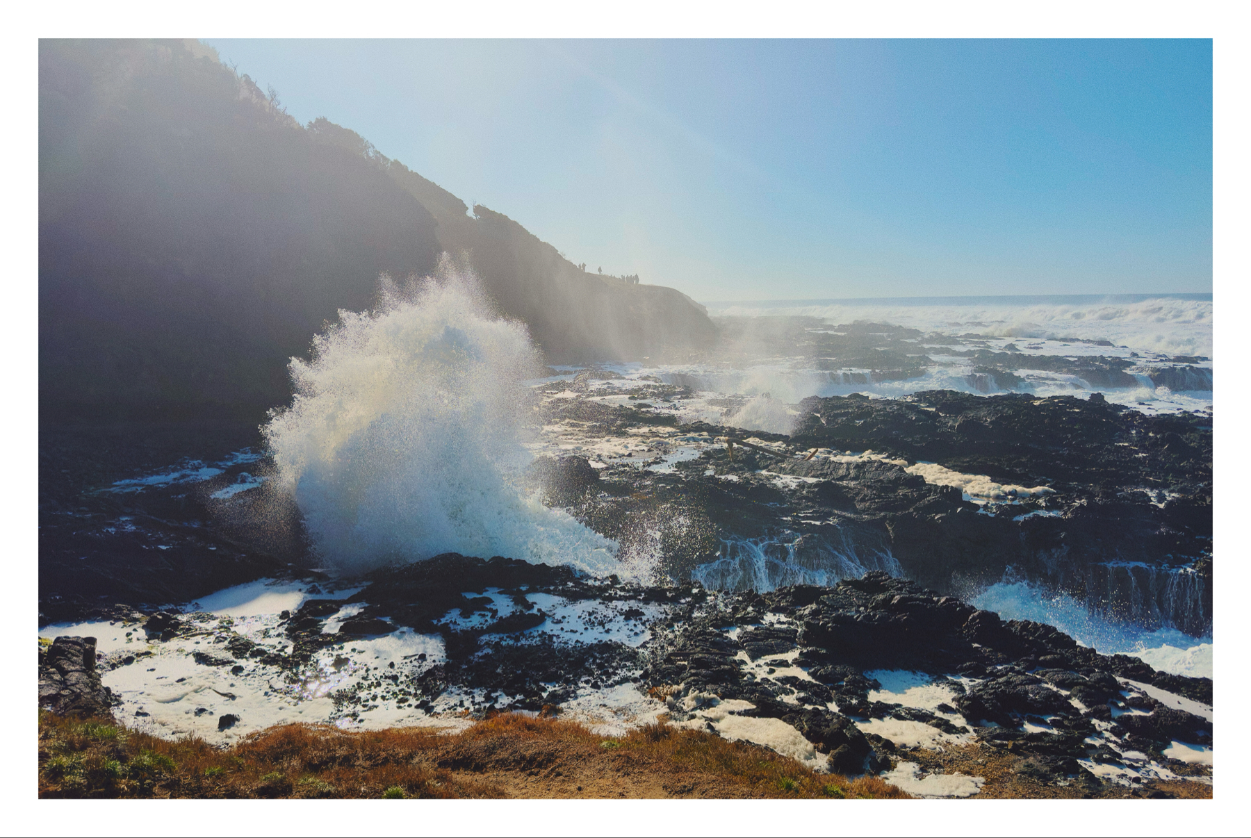 Coastal landscape with waves crashing against rocky shoreline, generating a large spray, under a clear sky with sunlight.