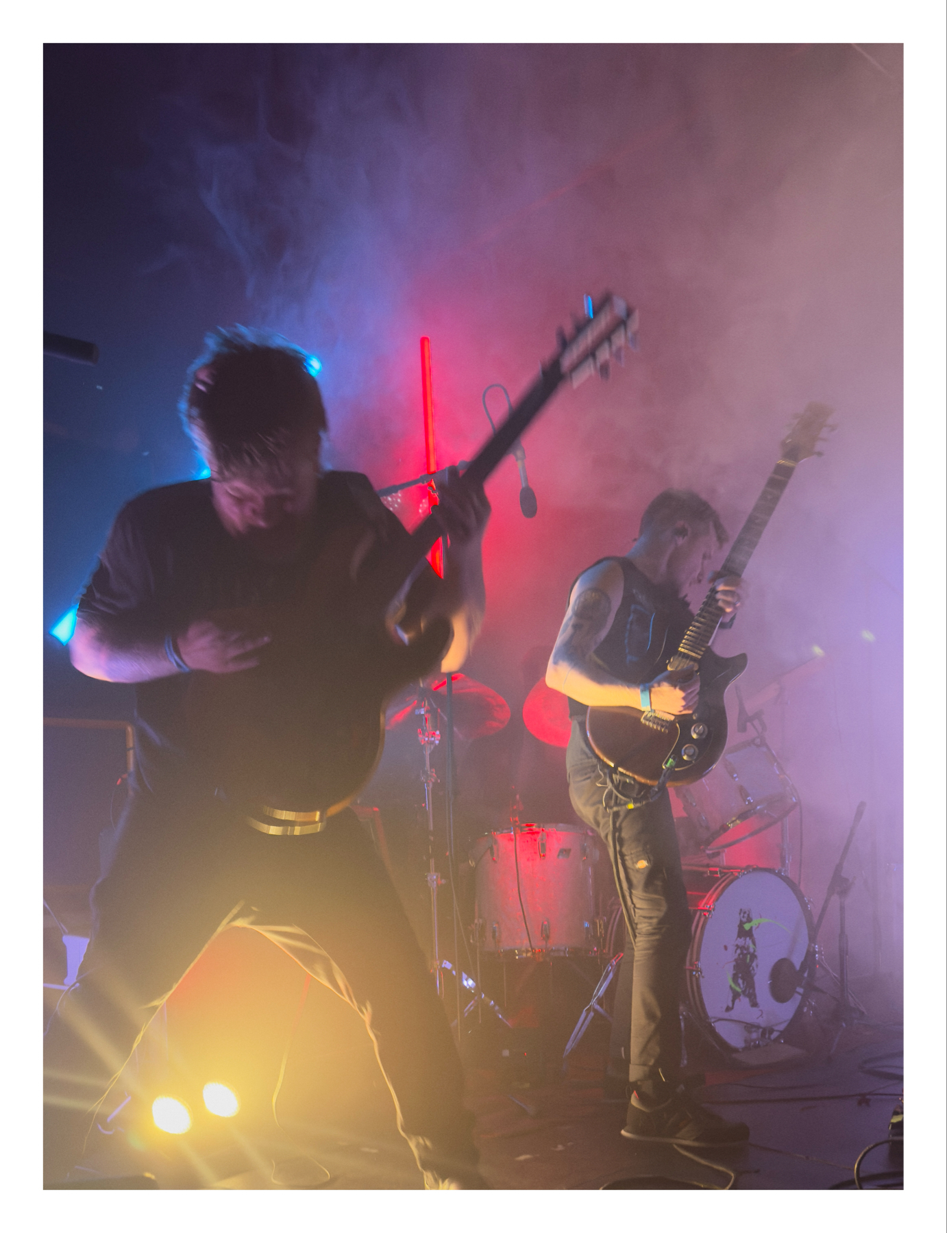 Two musicians from Shy, Low performing on stage, one singing and one playing an electric guitar, with dynamic lighting and smoke effects in the background.