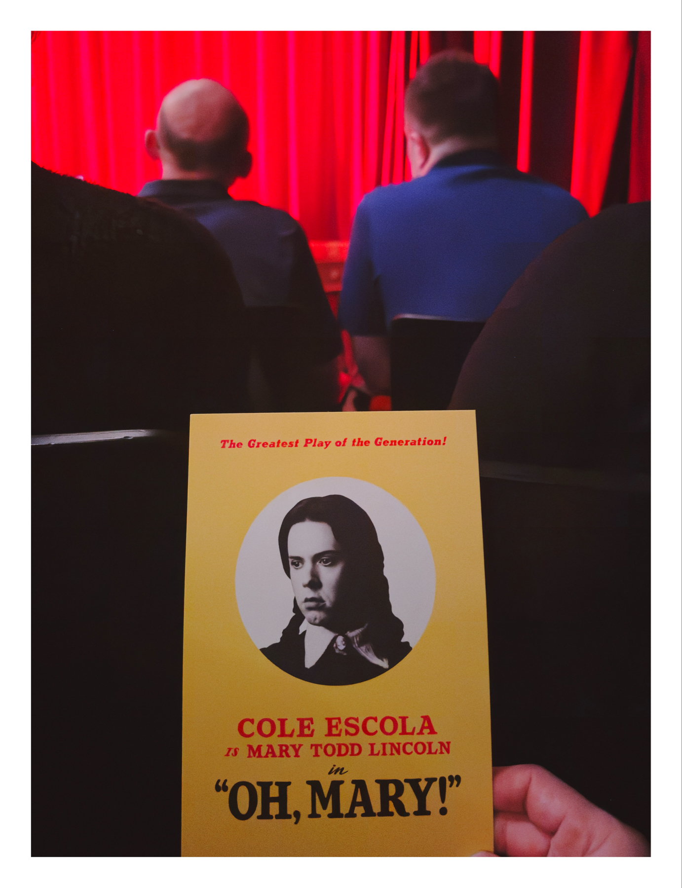 A theatre program for a play titled “OH, MARY!” featuring Cole Escola as Mary Todd Lincoln, held up against a blurred background featuring an audience waiting, with a red stage curtain in the distance.