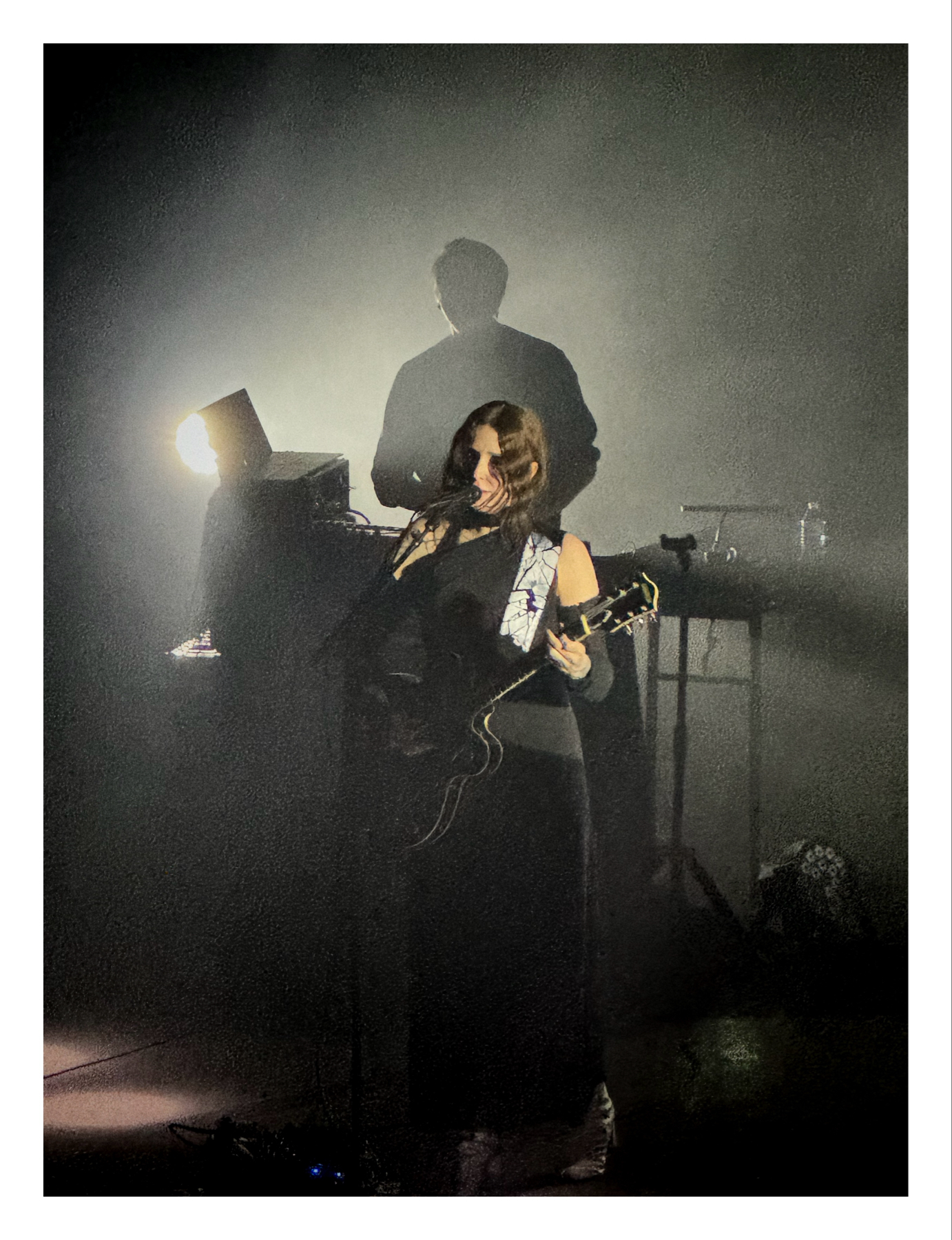 Chelsea Wolfe playing an electric guitar on stage with moody stage lighting casting their shadow on the wall behind.