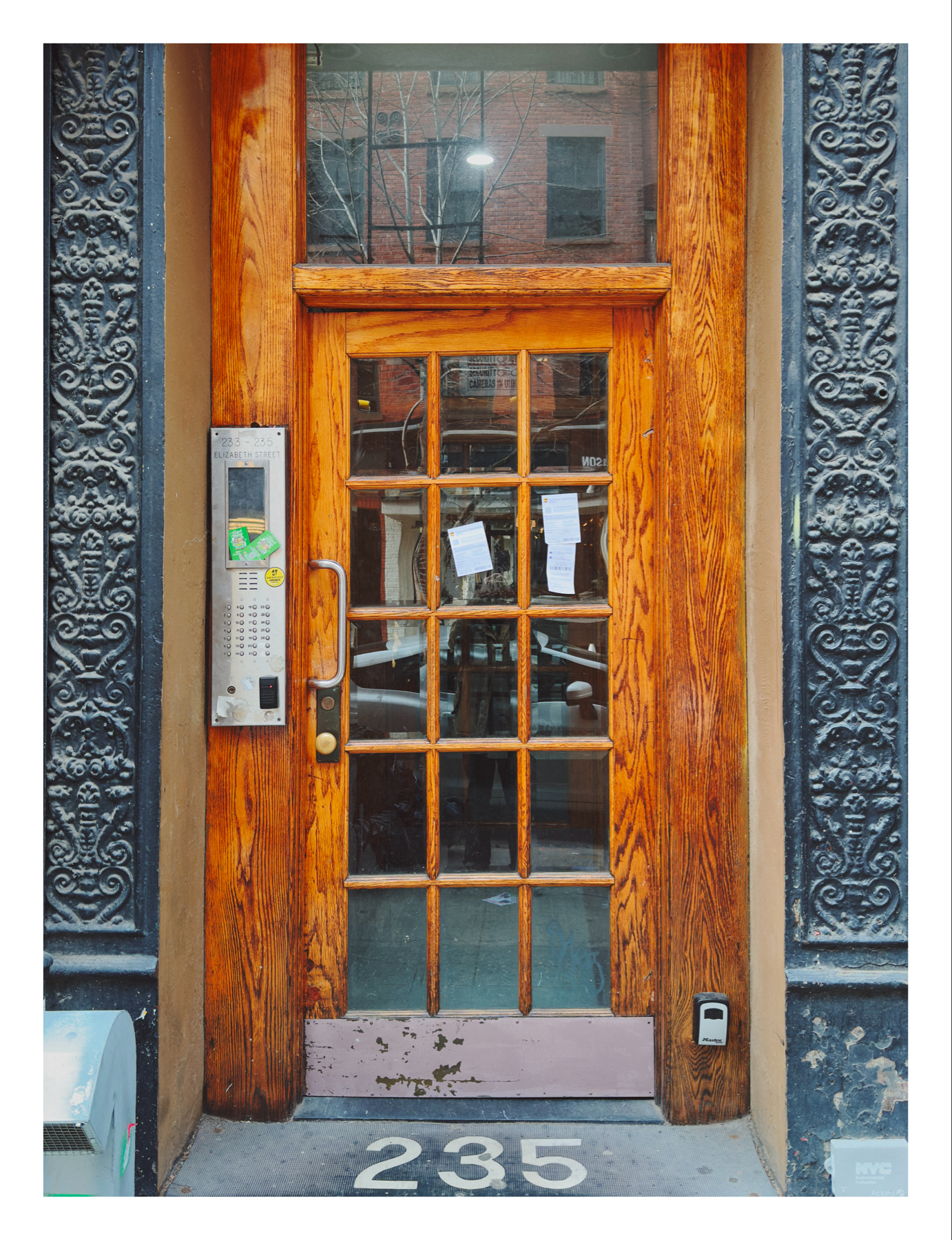 A wooden door with glass panes, framed by ornate metalwork, marked with the number 235. An intercom and buzzers are on the left, a small white device is to the right, and notices are attached to the door.
