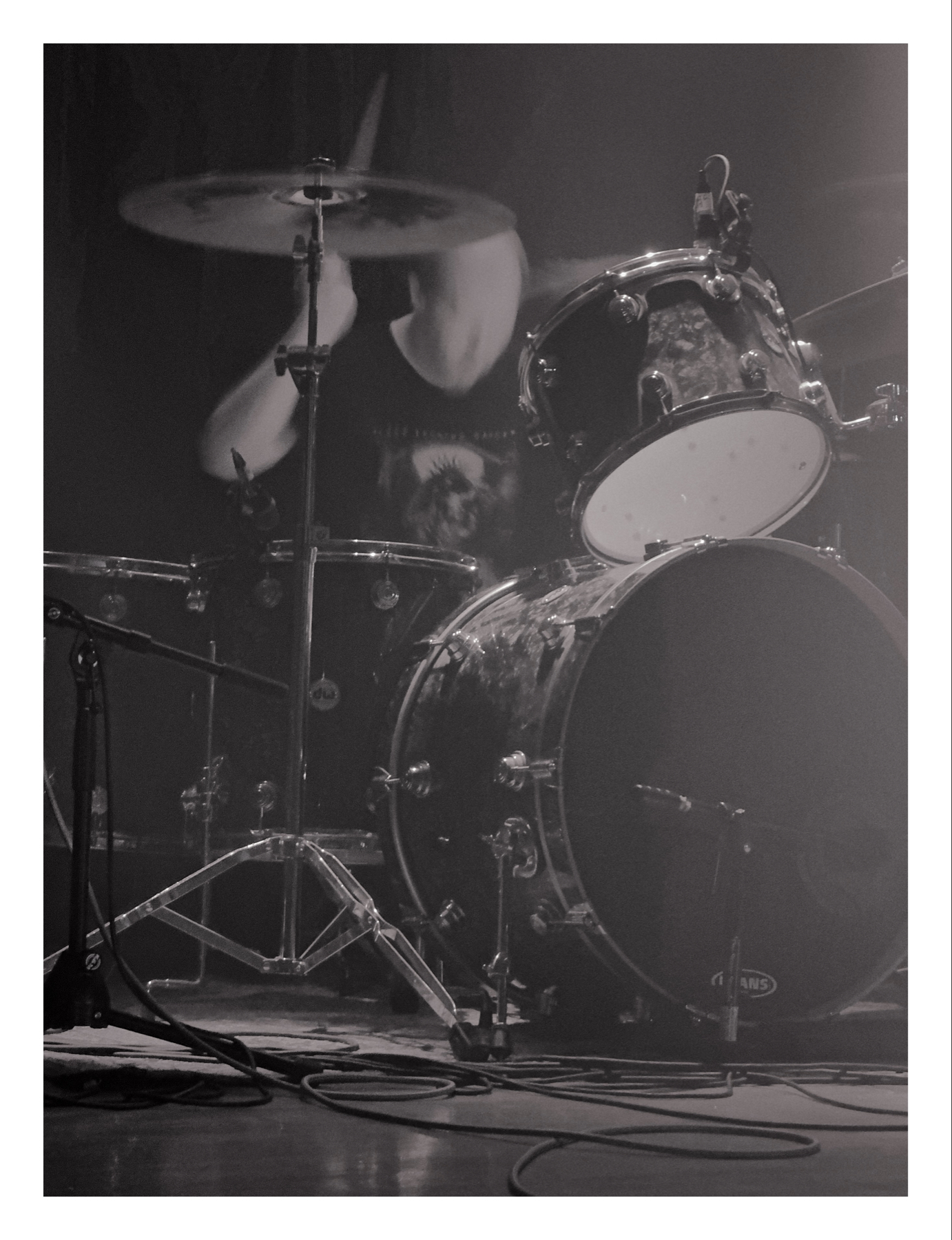 A black and white photo of a drum set on stage with a blurred drummer in motion behind the cymbals.