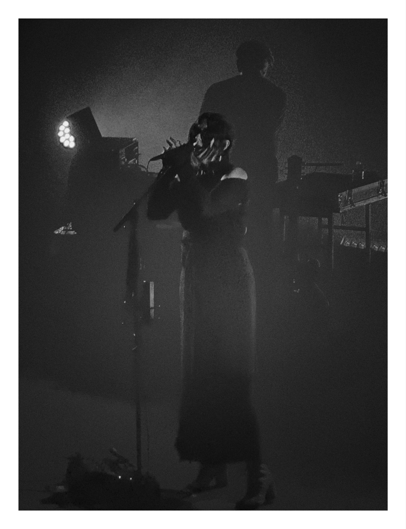 A black and white photo of Chelsea Wolfe singing into a microphone on a stage with dim lighting, with another person visible in the background.