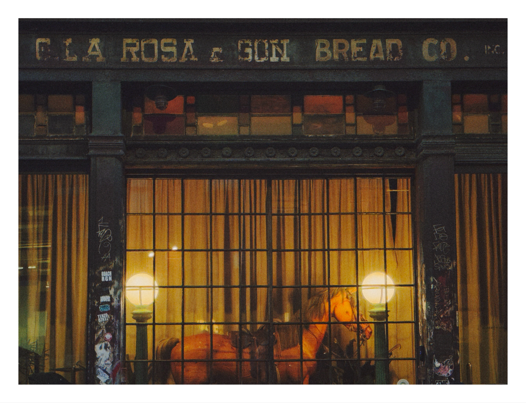 A storefront at night with illuminated windows, showcasing the name “C.I.A. ROSA & SON BREAD CO.” above. Warm light spills from the interior onto the sidewalk, casting a glow on urban elements like graffiti and street posters. There’s a horse inside the window. 