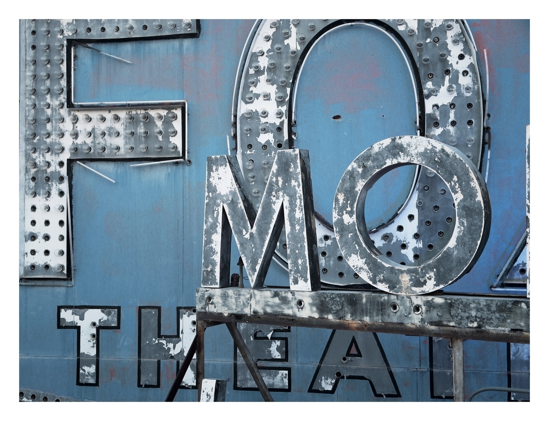 Weathered metal letters on a faded blue background possibly from a sign, exhibiting signs of age and disrepair.