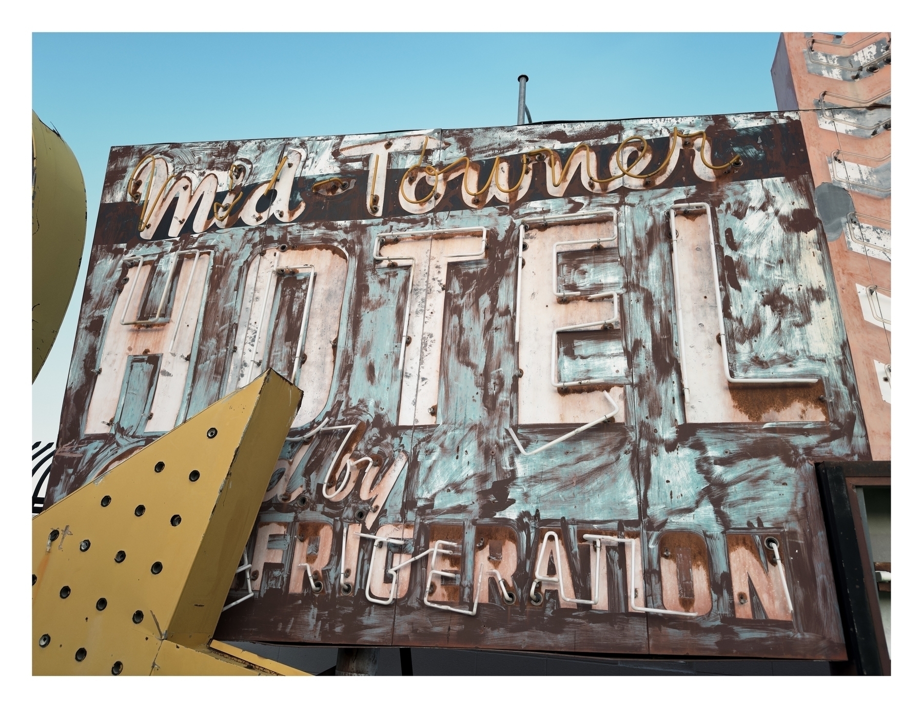 A weathered hotel sign reads “Midtown HOTEL by REFRIGERATION” with faded and rusty letters against the sky.