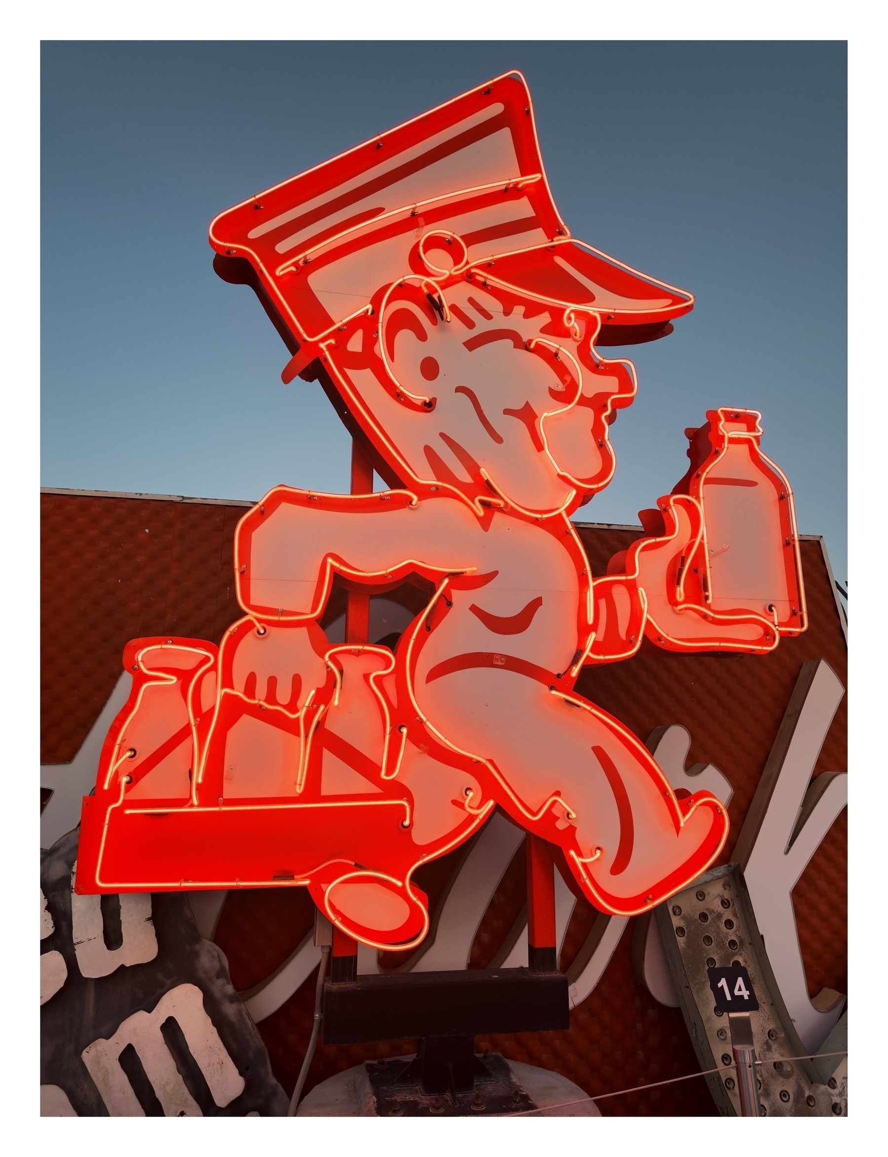 Red neon sign depicting a cartoon figure in a running pose, carrying a bottle, against a clear sky; mounted on a metal structure.