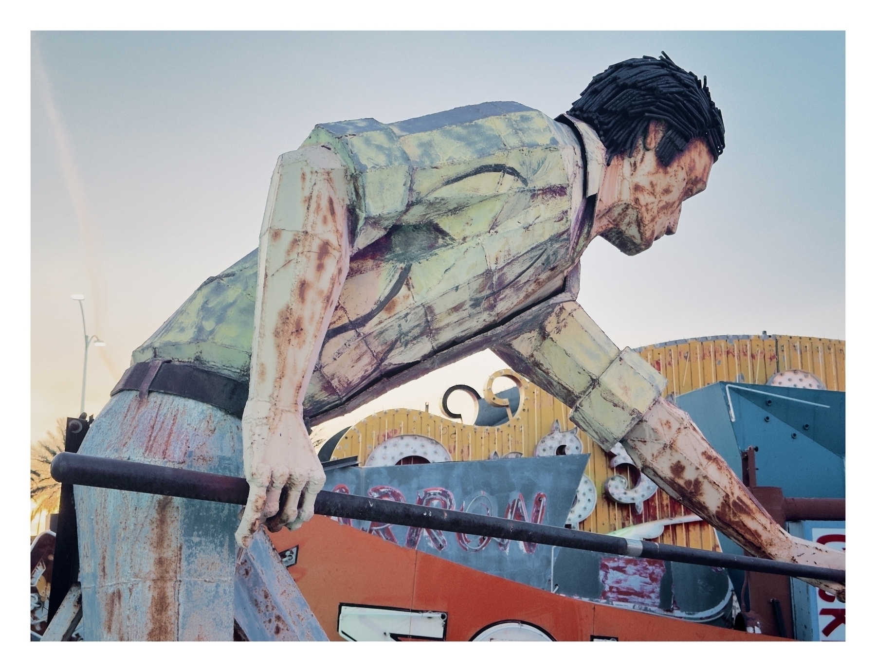 A large, weathered statue of a man leaning over a railing against a background of a colorful, cluttered scrapyard.